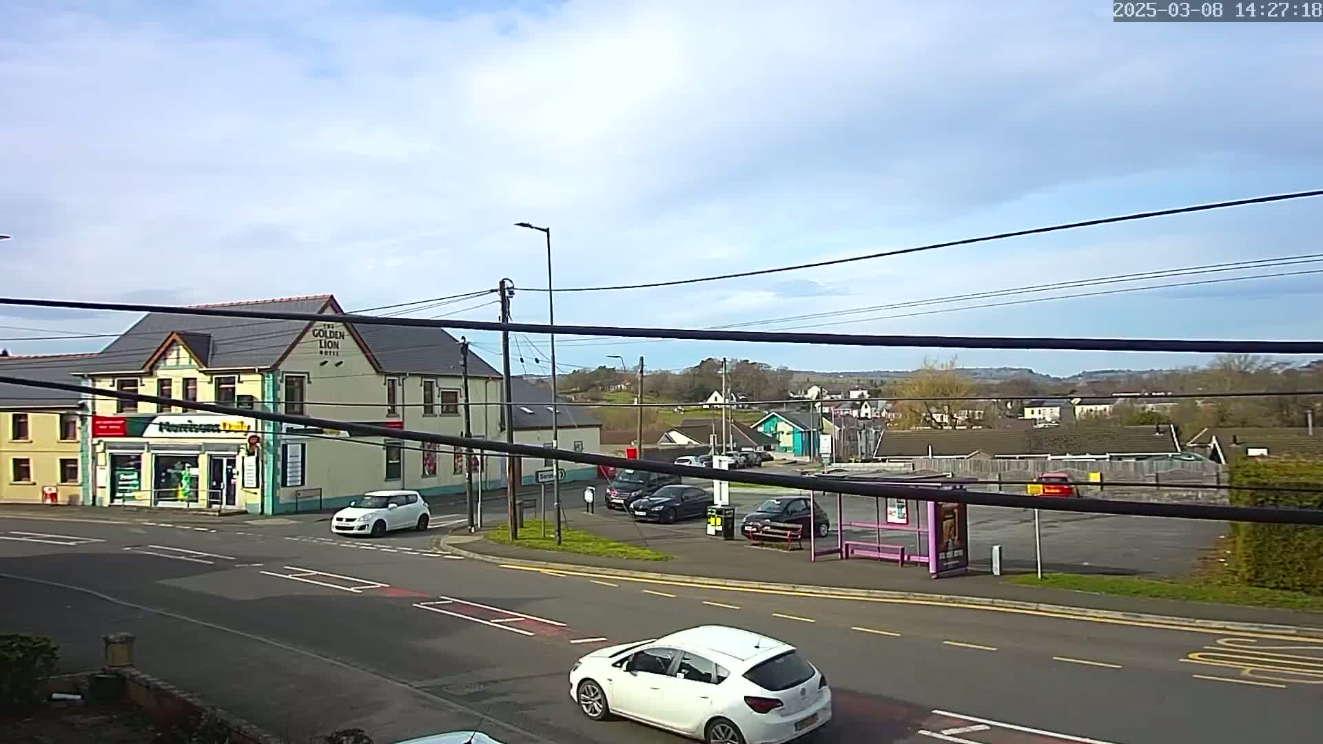A mostly sunny day reveals a street scene with several cars, a two-story building, a bus shelter, and a distant view of hills and houses.