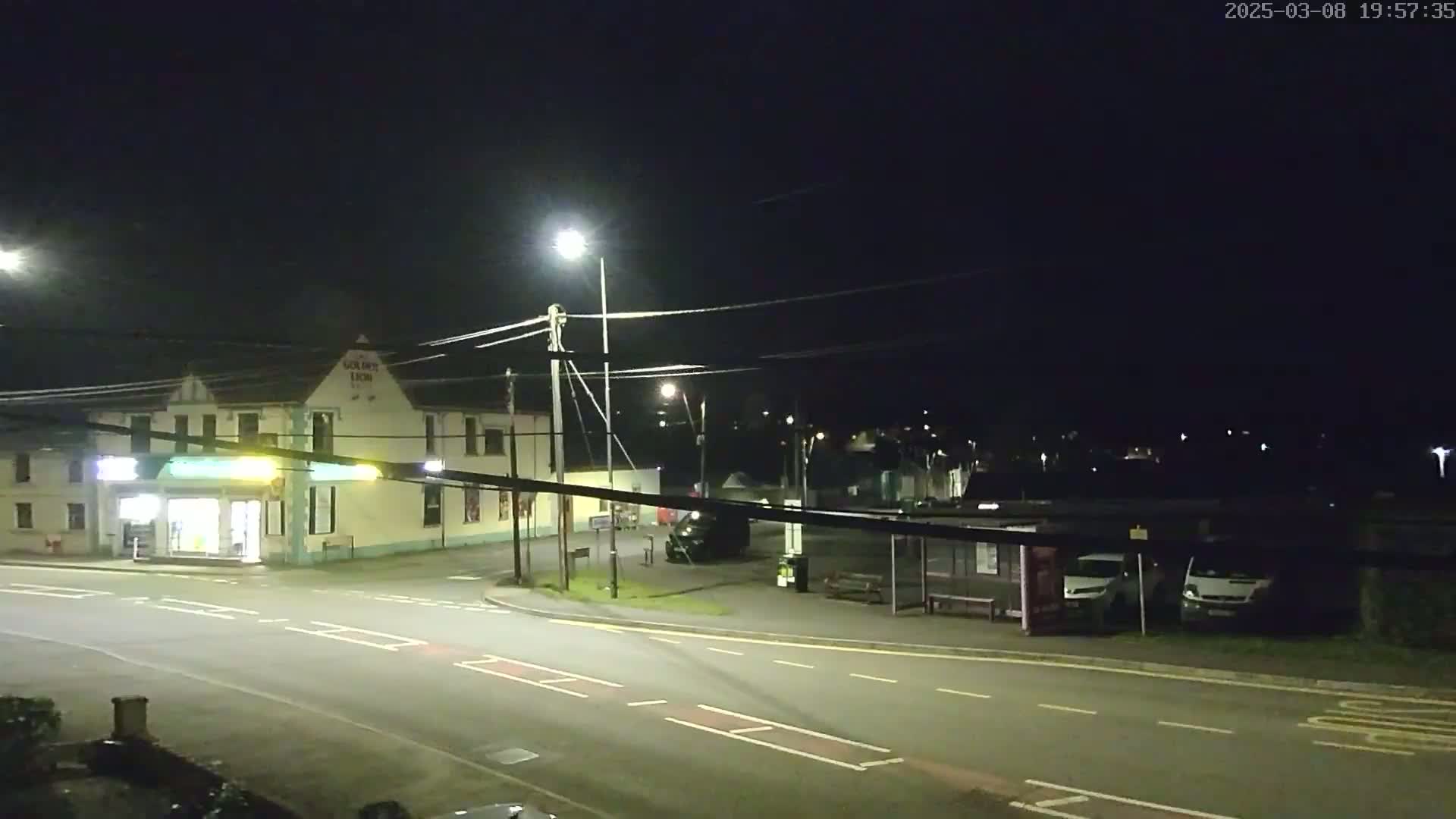 A nighttime street scene shows a two-story building with a shop on the ground floor, a bus stop with a few vehicles, and a road intersection under artificial light.