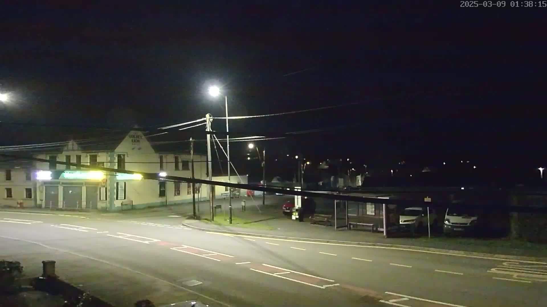 A nighttime view of a street intersection with a building, streetlights, parked vehicles, and a bus stop under a dark sky.