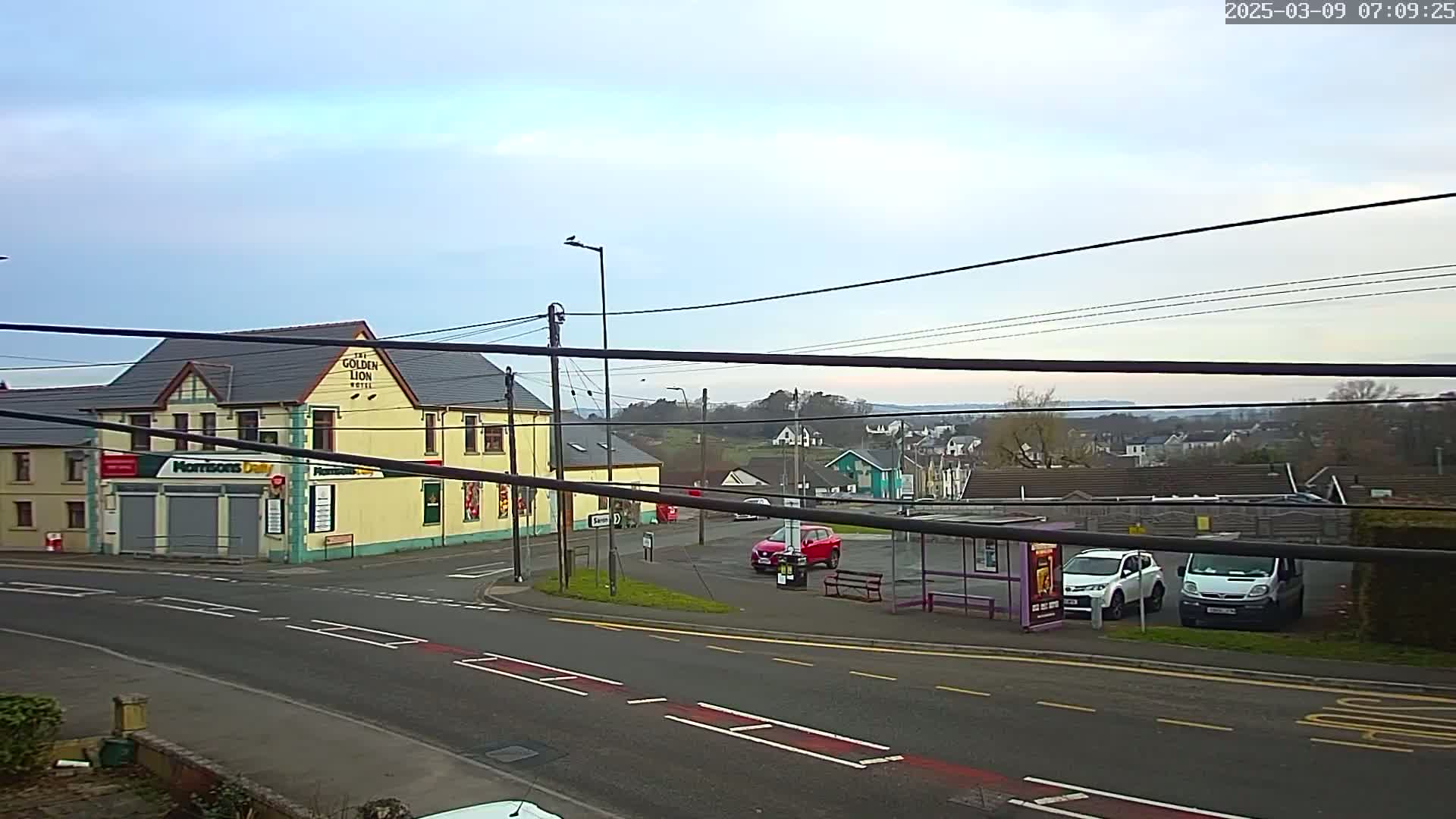 A mostly cloudy day reveals a street scene with a yellow building, several parked cars, a bus stop, and a view of houses on a distant hill.
