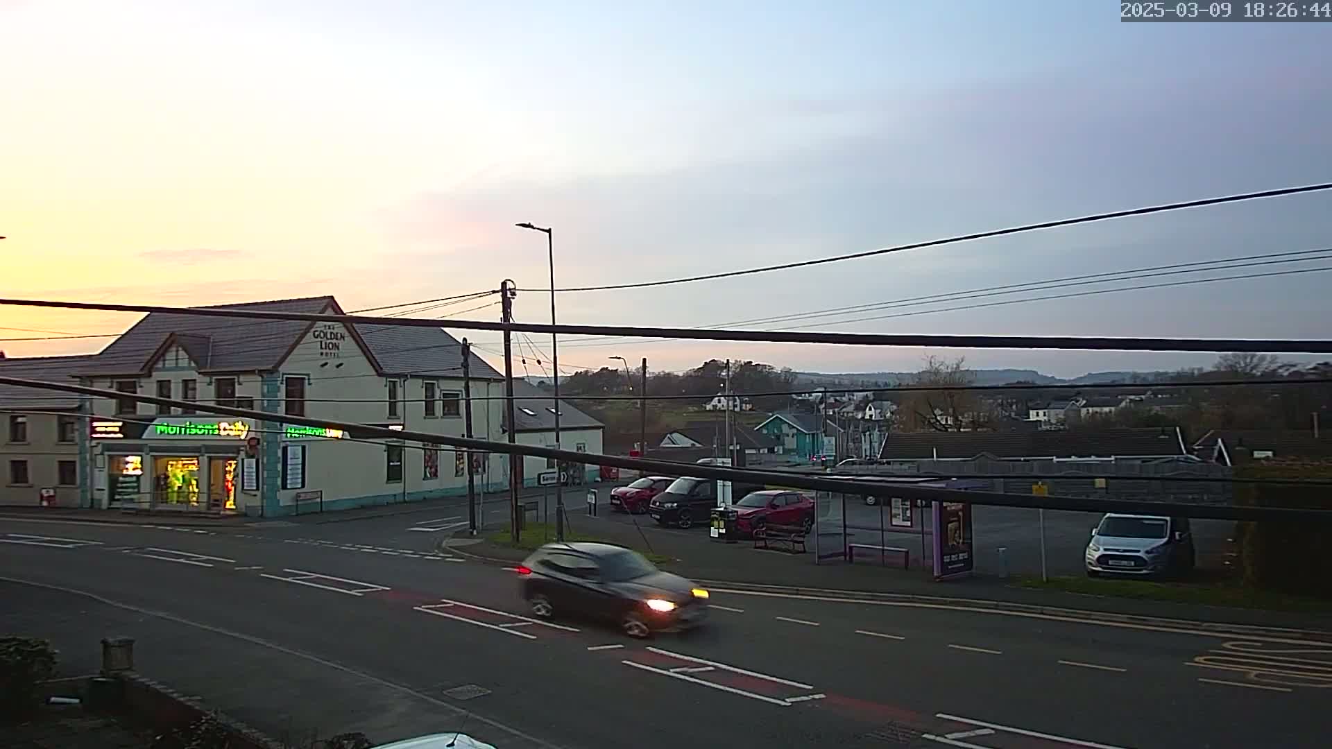 A twilight scene shows a street corner with a pub and a small shop, several cars parked and driving by, and a bus shelter in a parking lot, all under a mostly clear sky.