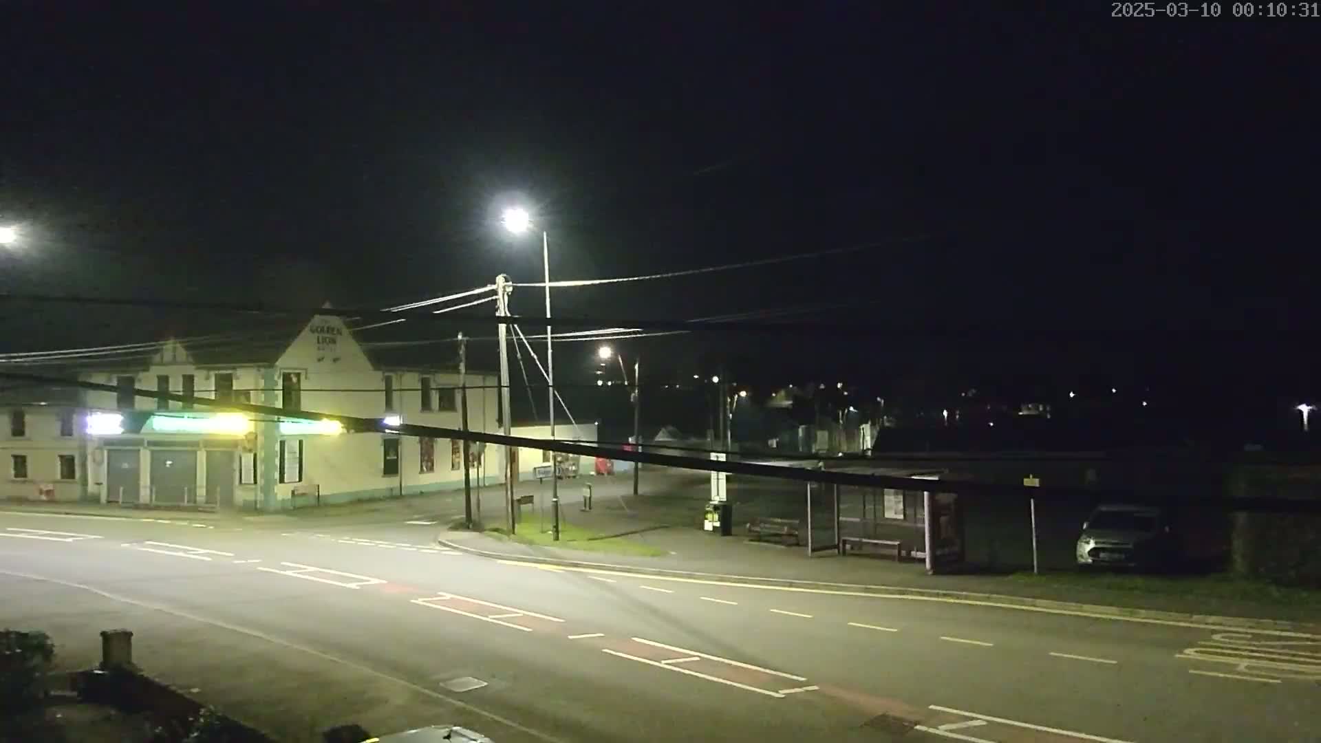 A nighttime street scene shows a building with illuminated signage, streetlights, a bus stop with a vehicle parked nearby, and a sparsely populated road intersection.