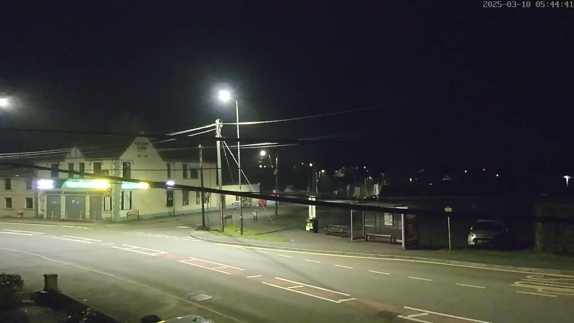 A nighttime street scene shows a building with illuminated signage, a bus shelter, a parked car, and streetlights under a clear, dark sky.