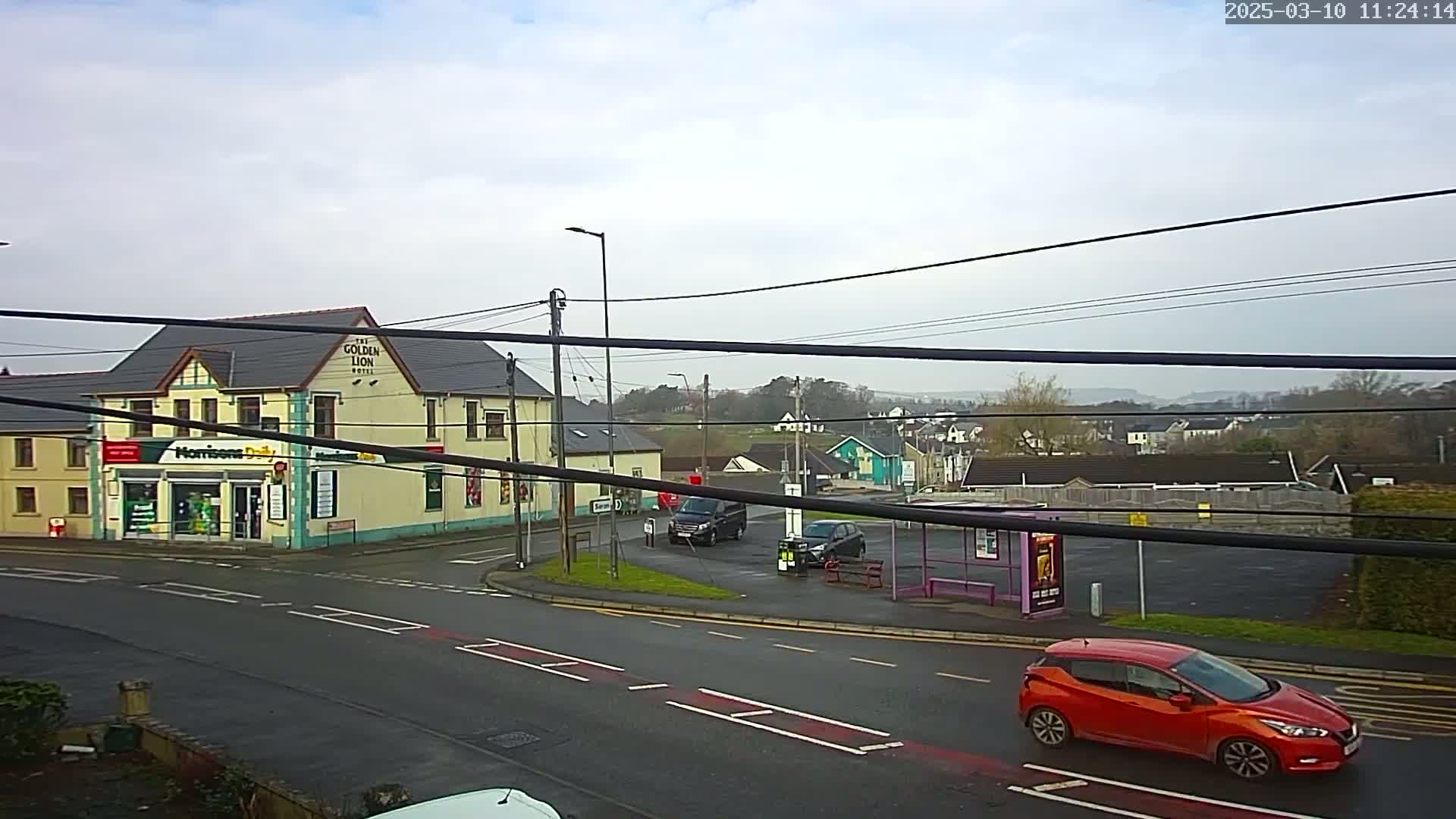 A street corner scene on an overcast day shows a yellow building, a small shop, several cars, a bus stop, and a residential area in the background.
