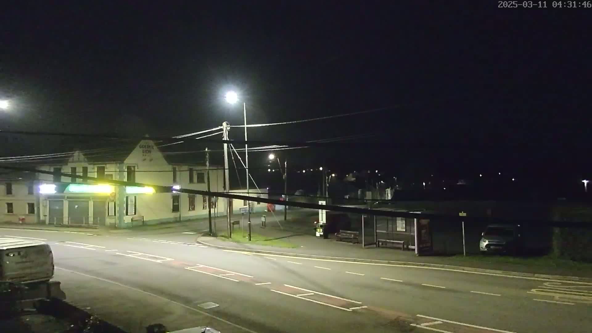 A nighttime street scene shows a light-colored building, a bus shelter, a few parked cars, and a road intersection illuminated by streetlights under a dark sky.