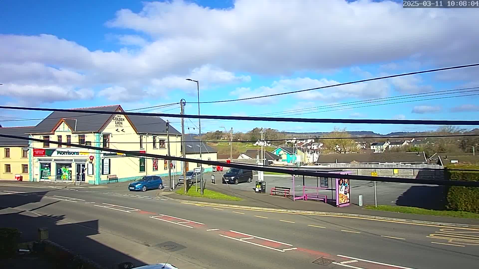 A sunny day with partly cloudy skies shows a street corner with a hotel, a convenience store, several vehicles, and a bus stop.