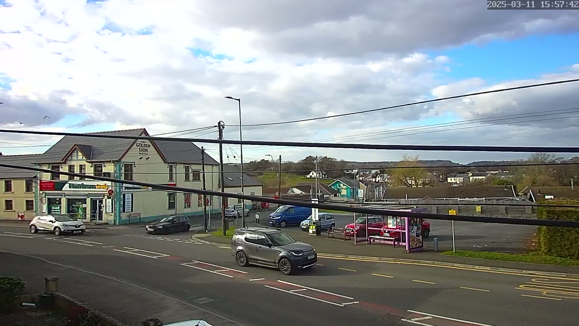 A partly cloudy day reveals a street scene with several cars, a bus stop, and a pub called "The Golden Lion Hotel," all situated near a small town or village.