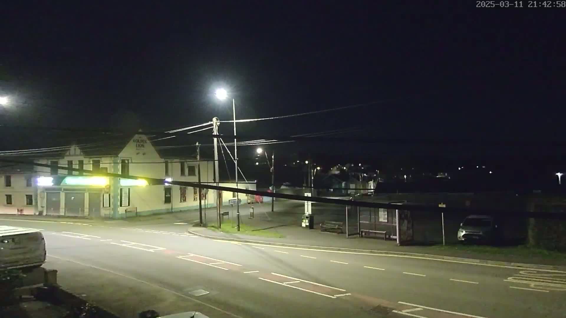 A nighttime street scene shows a building with lights on, a bus stop, and a few parked vehicles under artificial streetlights.
