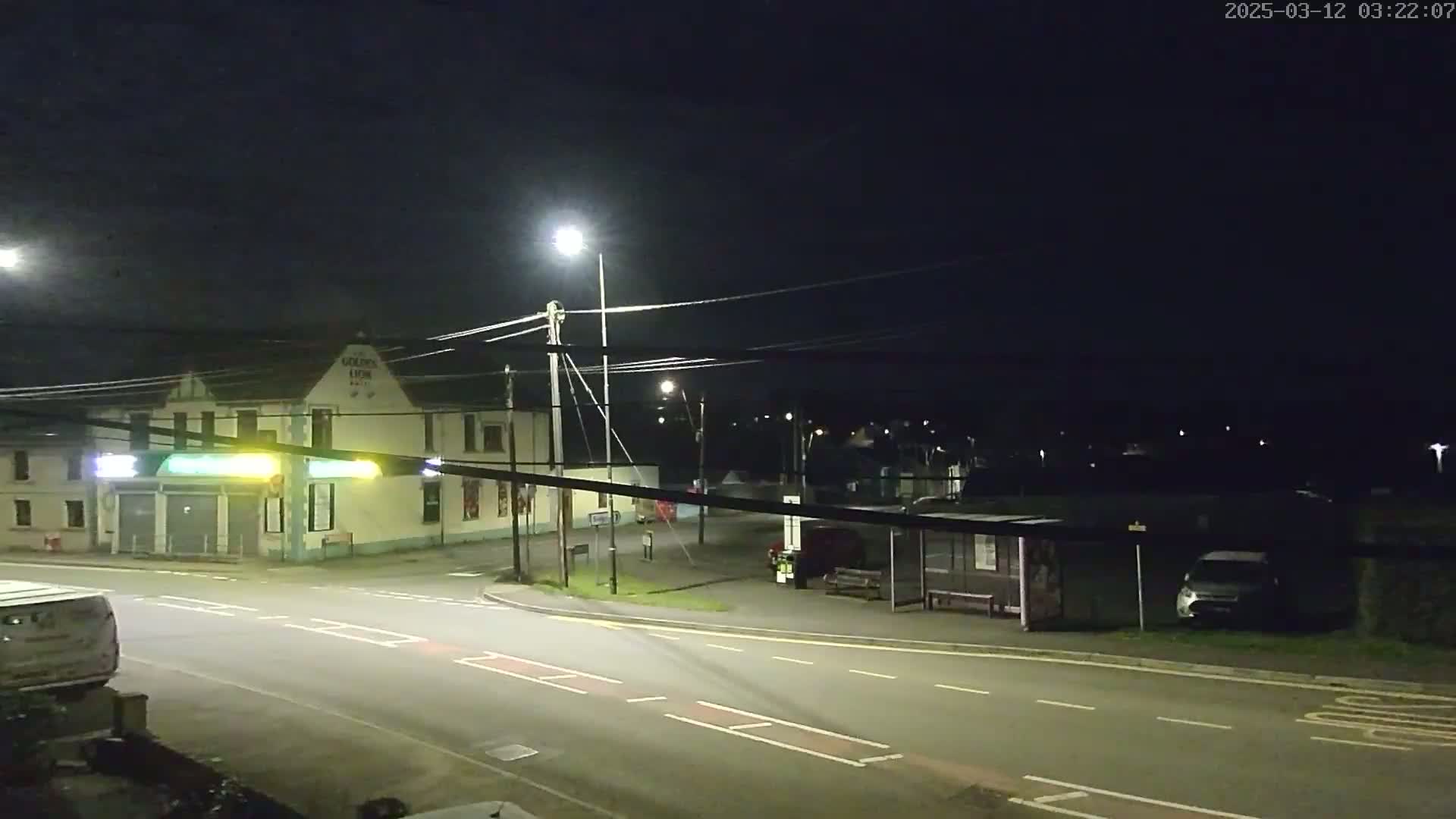 A nighttime street scene shows a light-colored building, a bus shelter, and several cars parked near a road intersection under bright streetlights.