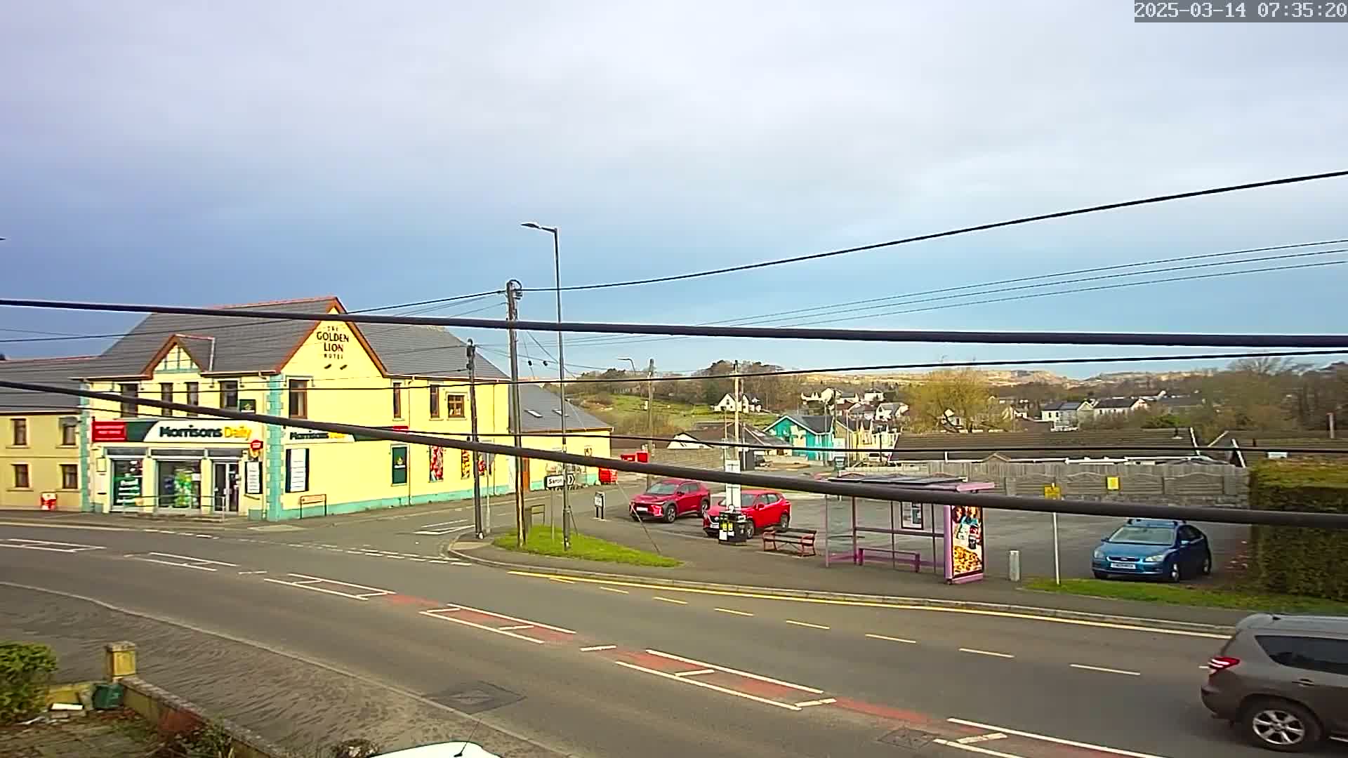 A partly cloudy day reveals a street scene with a yellow building containing a store and a pub, several cars parked near a bus stop, and a residential area visible in the background.