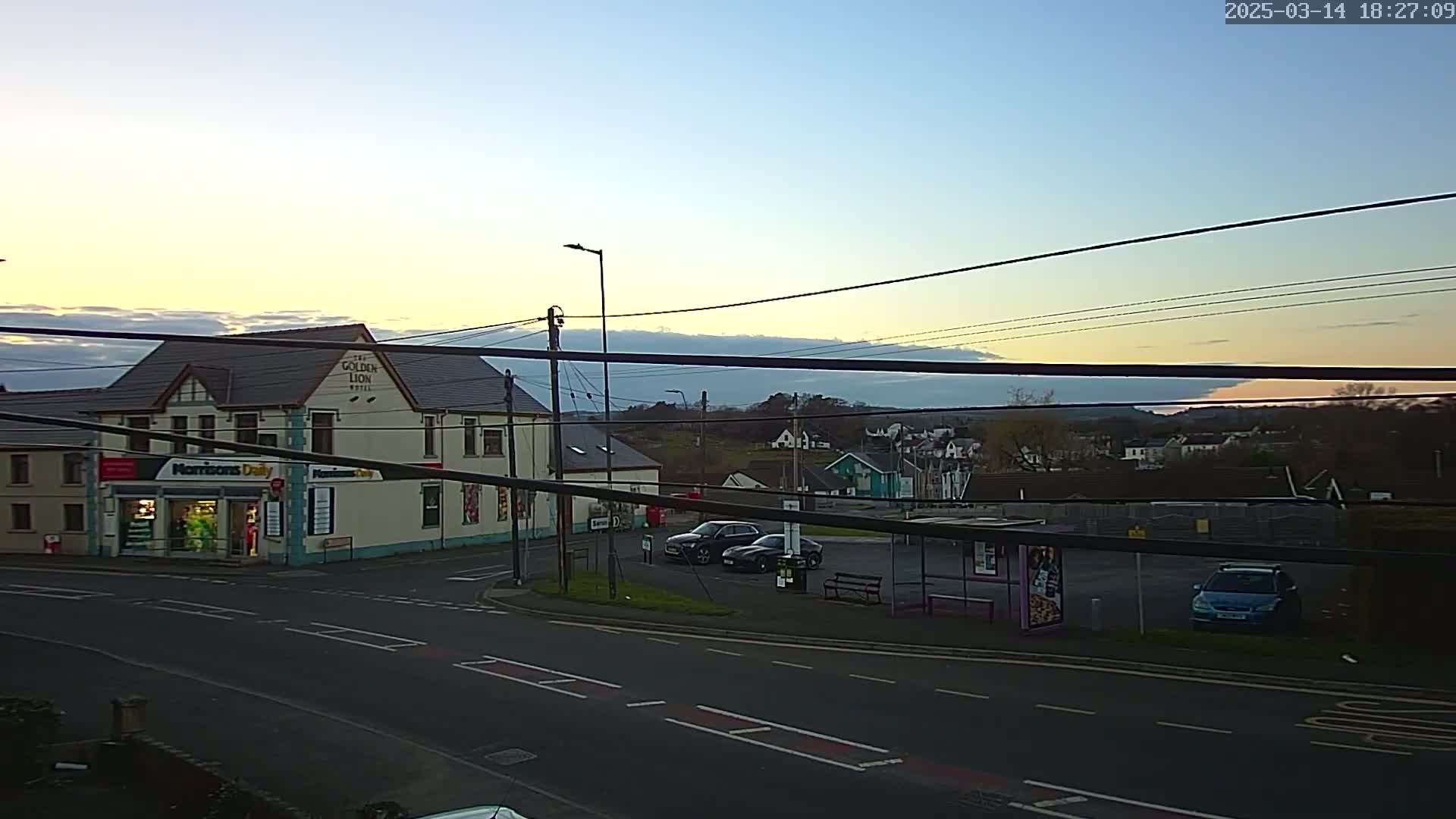 A street scene at twilight shows a two-story building with a shop on the ground floor, several cars parked in a lot across the street, and a bus shelter, all under a mostly clear sky with a thin band of clouds near the horizon.