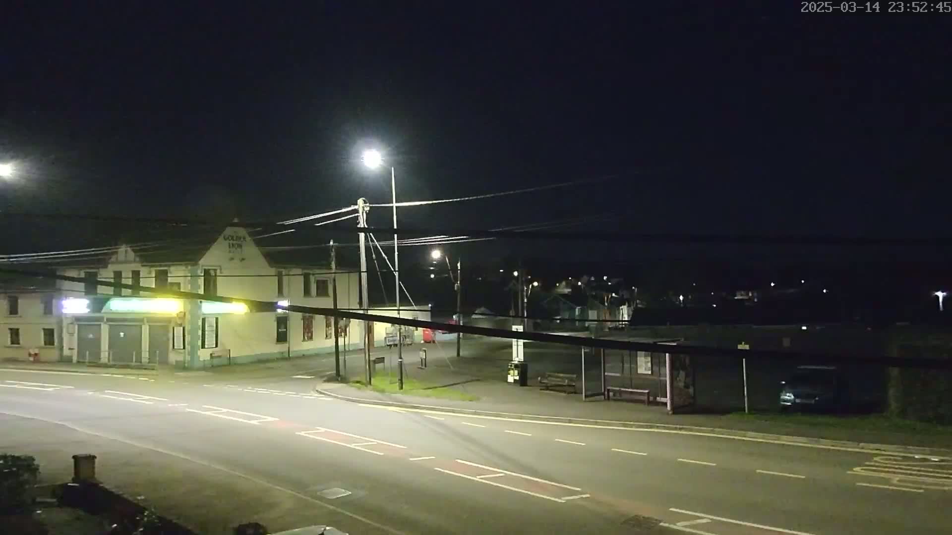 A nighttime street scene shows a building with a streetlight in front, a bus stop, and a few cars parked on a dark road.