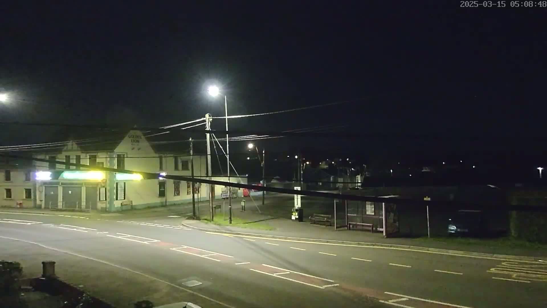 A nighttime street scene shows a building with lights on, a bus stop, and a mostly empty road under streetlights.
