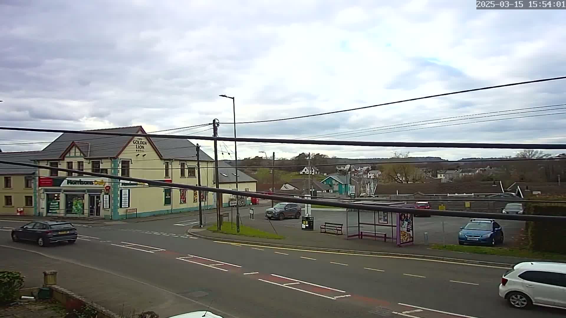 A street scene on an overcast day shows a corner with a pub, a small shop, a bus stop, and several cars parked in a lot.