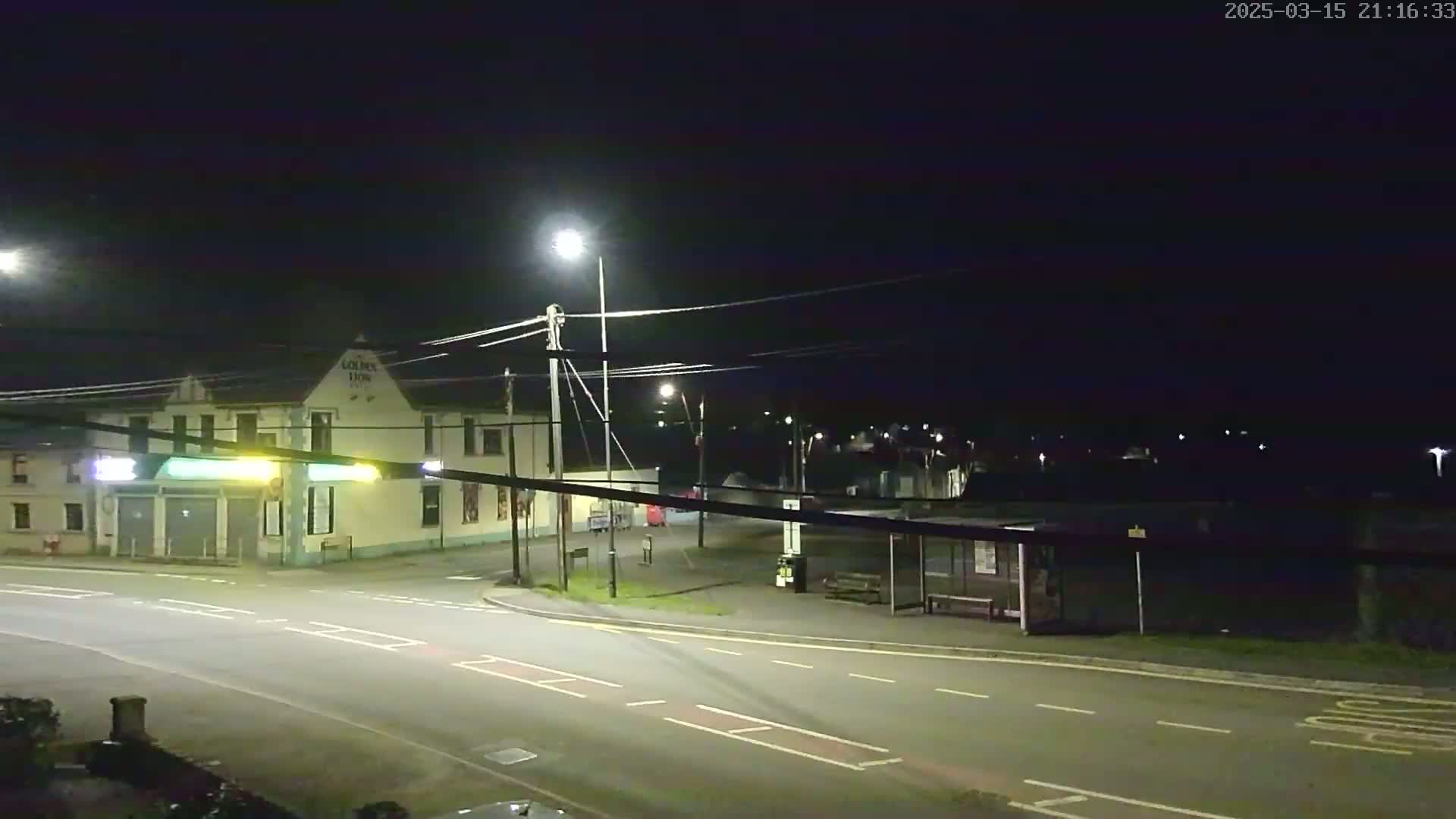 A nighttime street scene shows a two-story building with a bus stop across the street under illuminated streetlights.