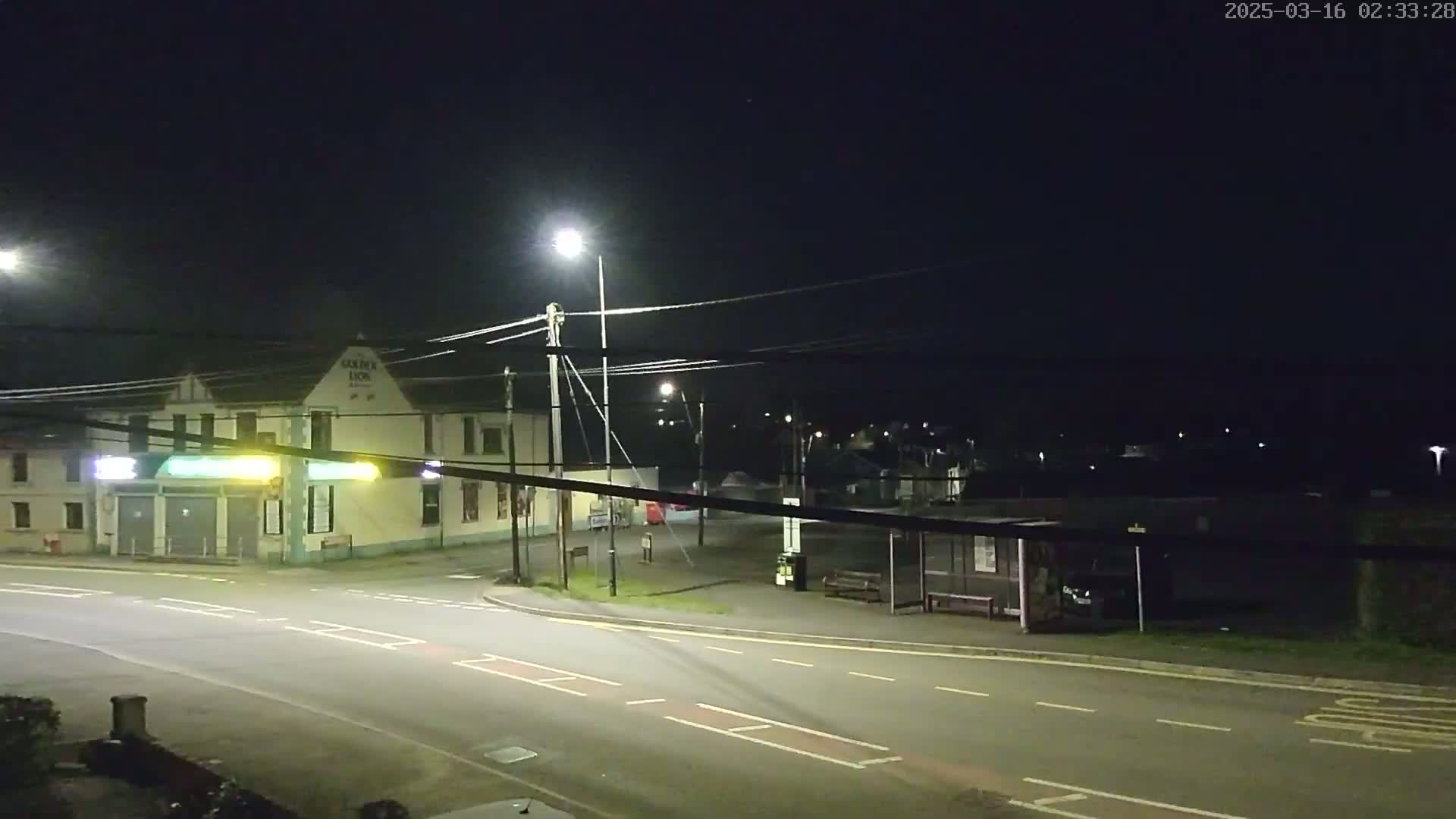 A nighttime street scene shows a building, a bus stop, and a road intersection under artificial light.