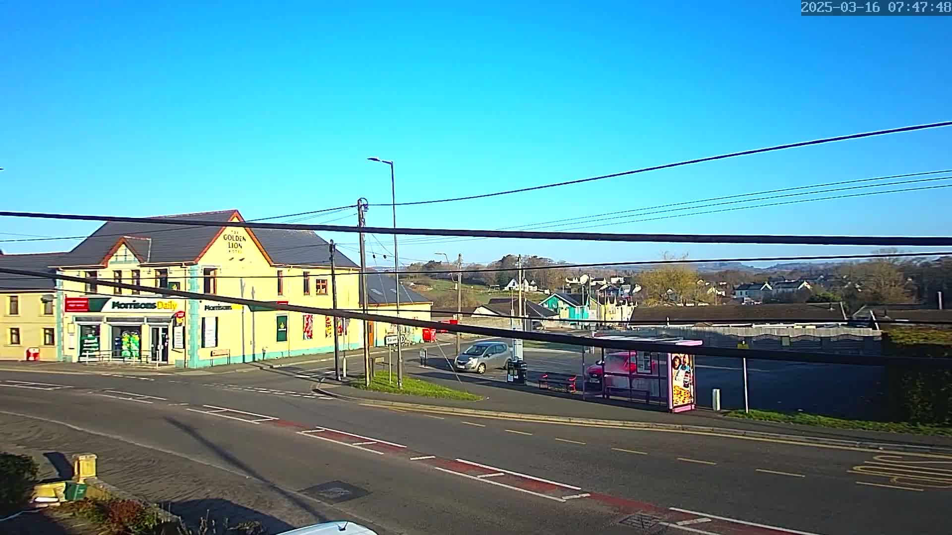 A yellow building with a store on the ground floor sits at a road intersection under a clear blue sky, with several cars parked nearby.