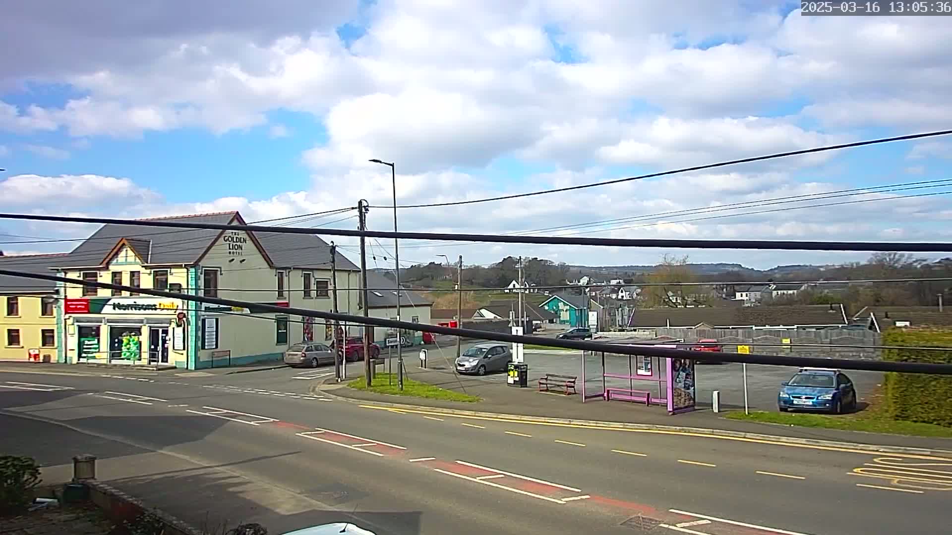 A partly cloudy day shows a street corner with a hotel, a small shop, several parked cars, a bus stop, and houses and hills in the distance.