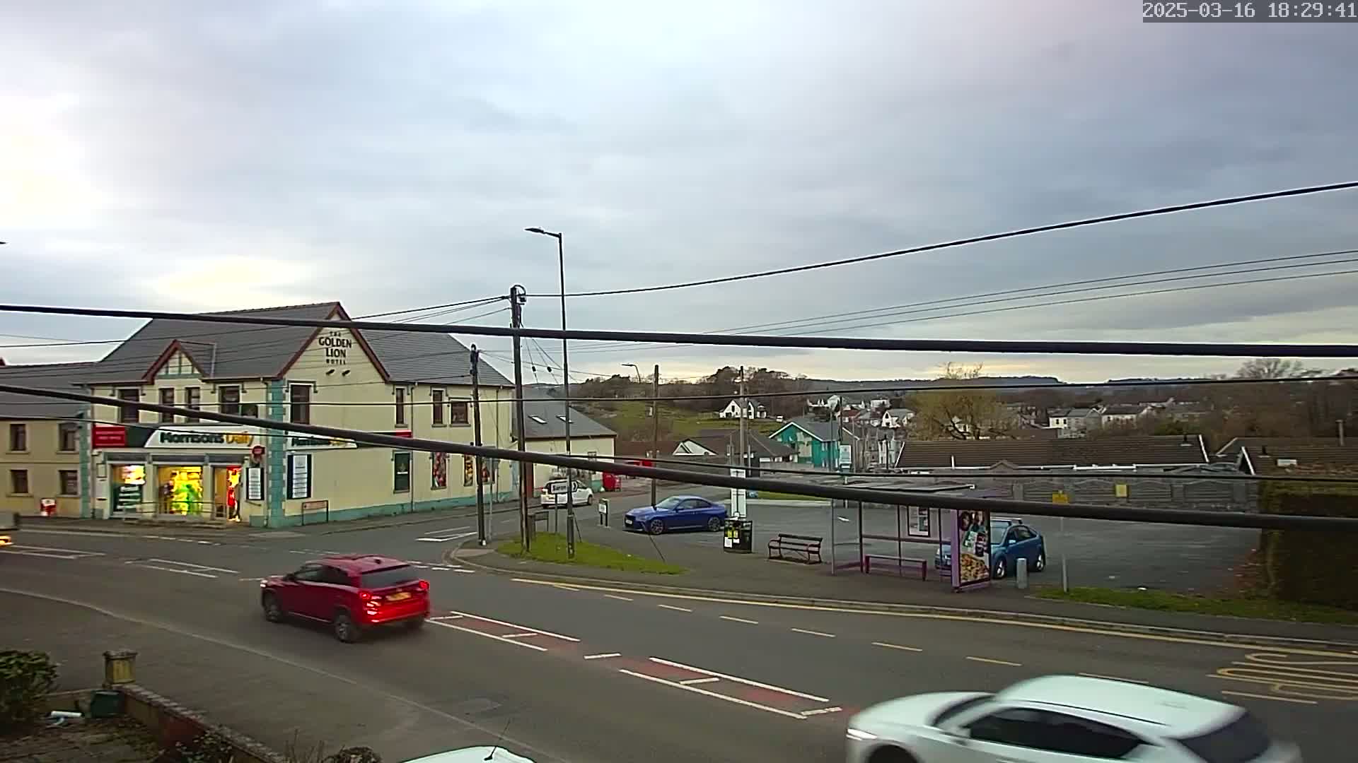 A street scene on an overcast day shows a yellow building with a shop and hotel, several cars, a bus stop, and houses on a hill in the background.