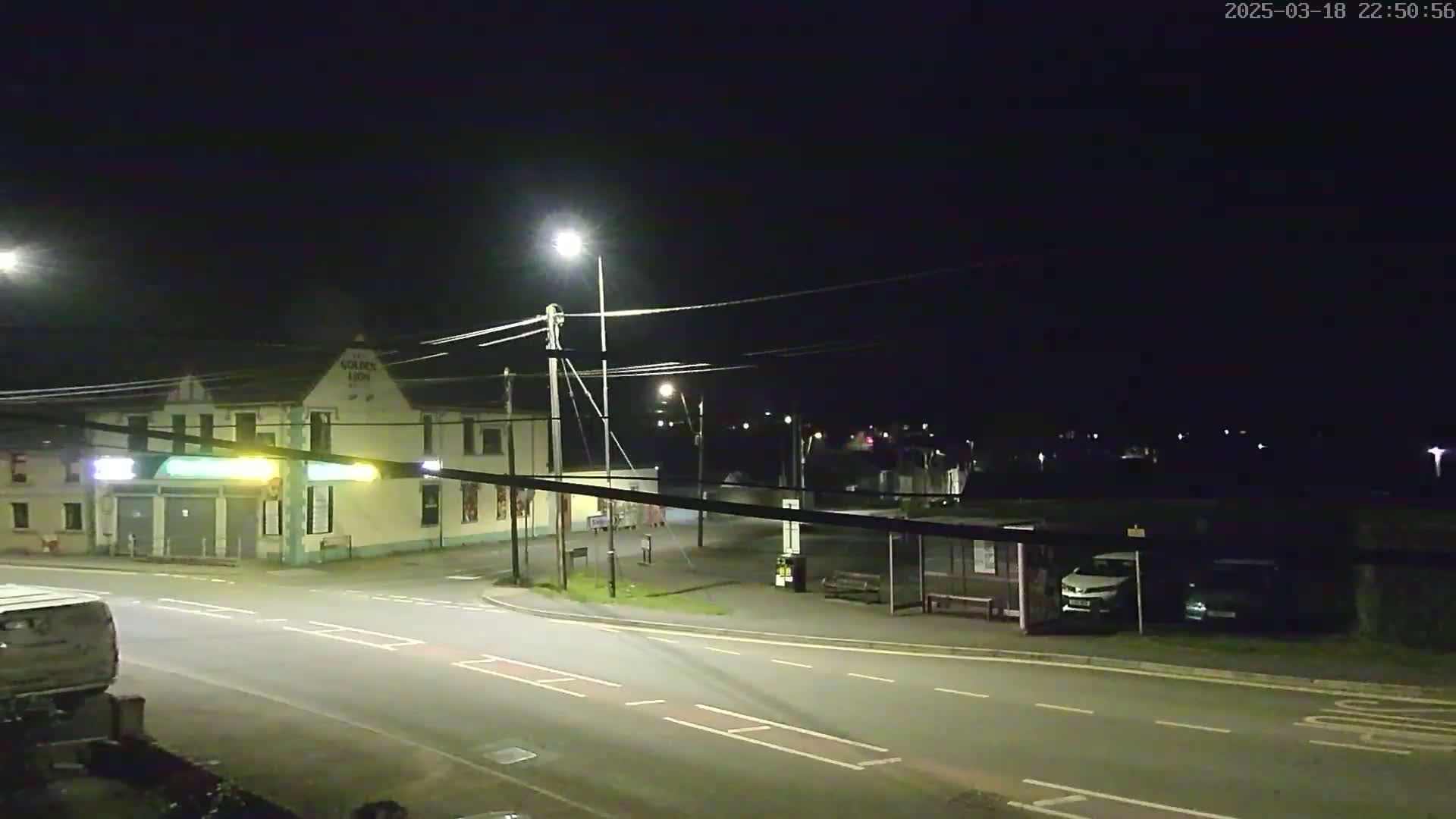 A nighttime street scene shows a building with streetlights illuminating a mostly empty road and a bus stop with a few parked cars.