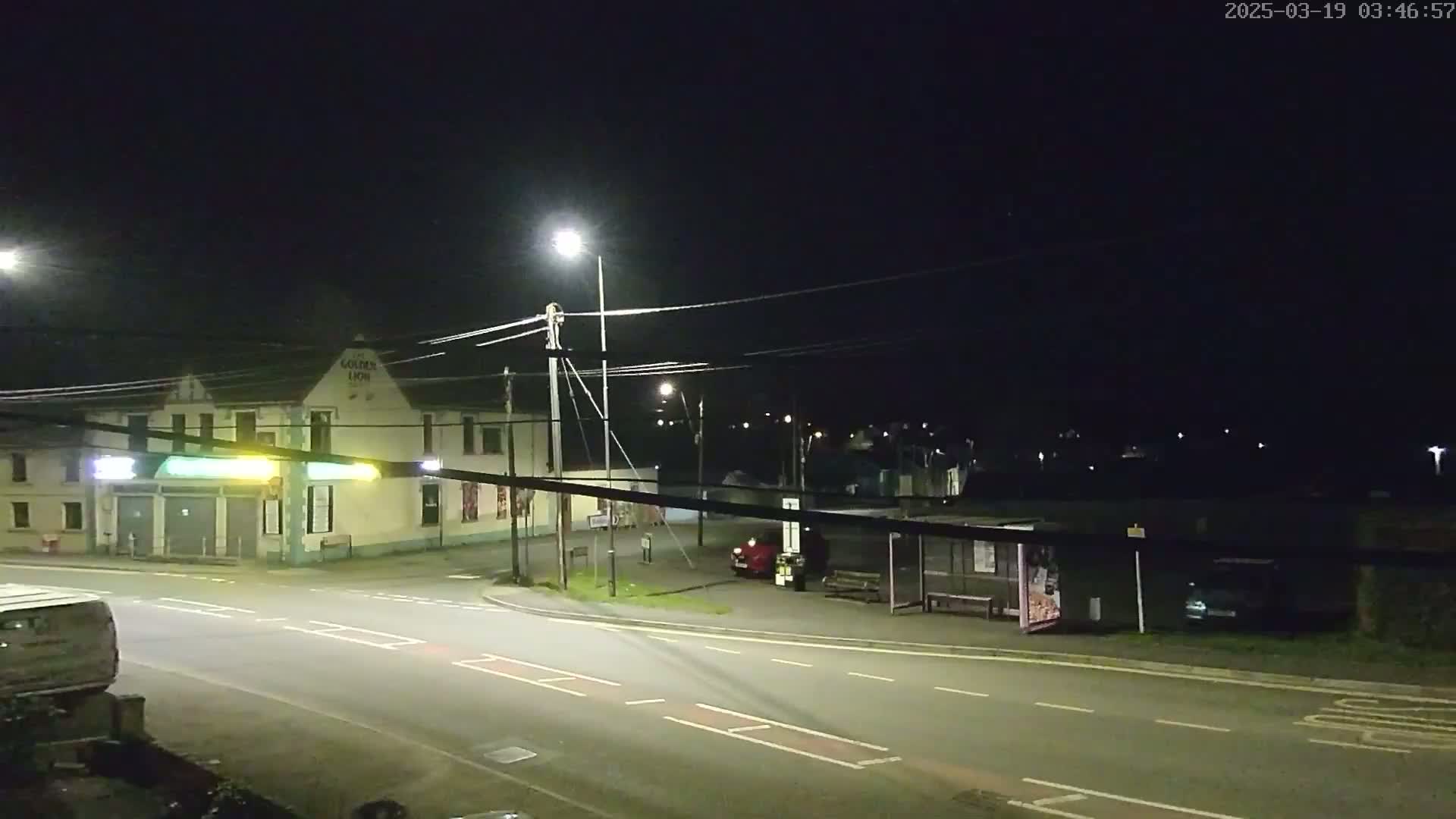 A nighttime street scene shows a building, a bus stop with a few vehicles parked nearby, and a roadway under artificial light.