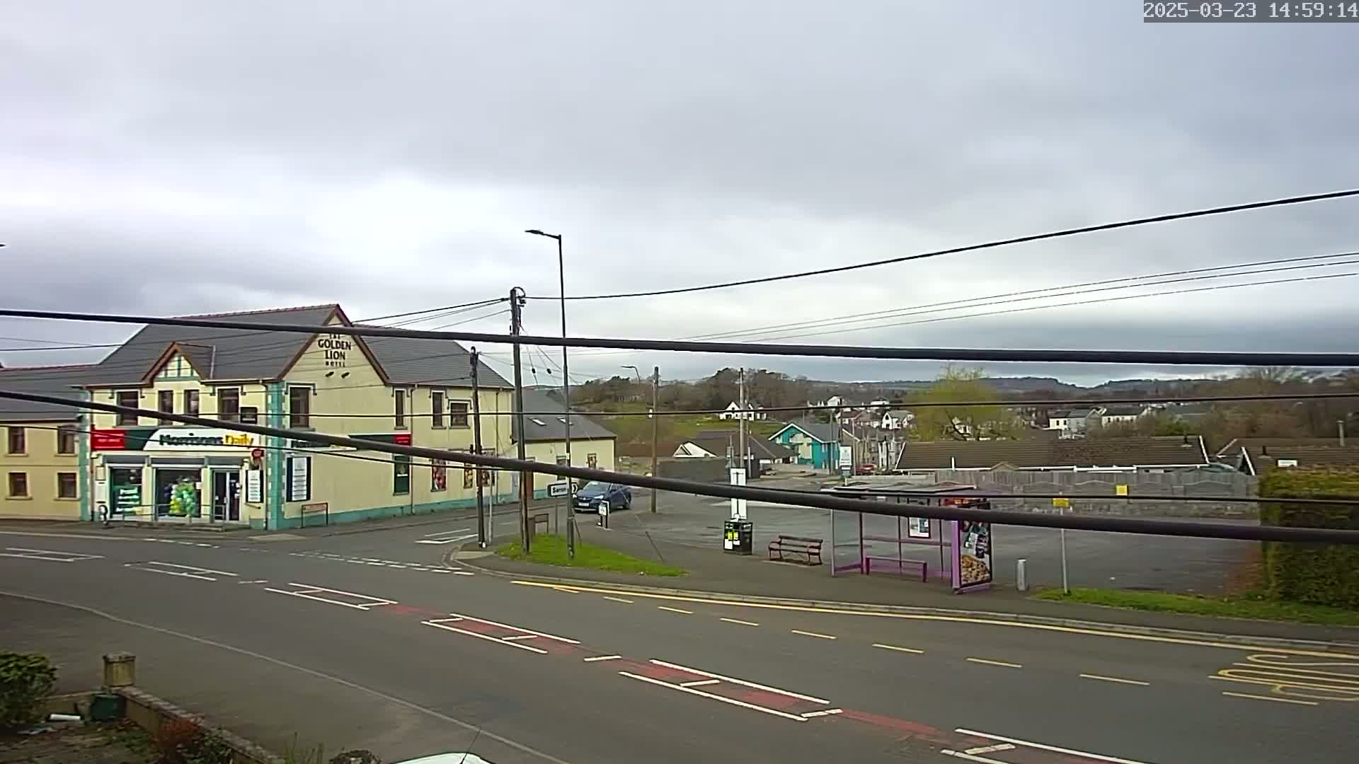 A street scene on an overcast day shows a building with a shop and a pub, a small parking lot with a bus shelter, and several cars.