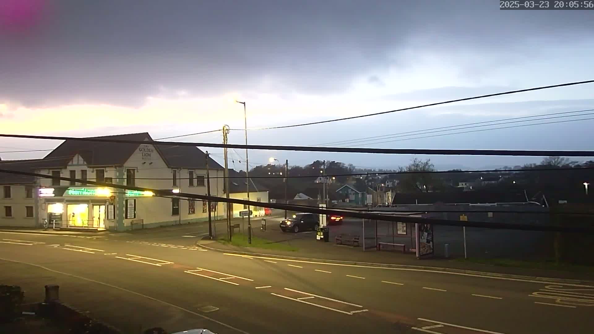 A dimly lit street scene at dusk shows a two-story building, a bus stop, and several cars under a cloudy sky.