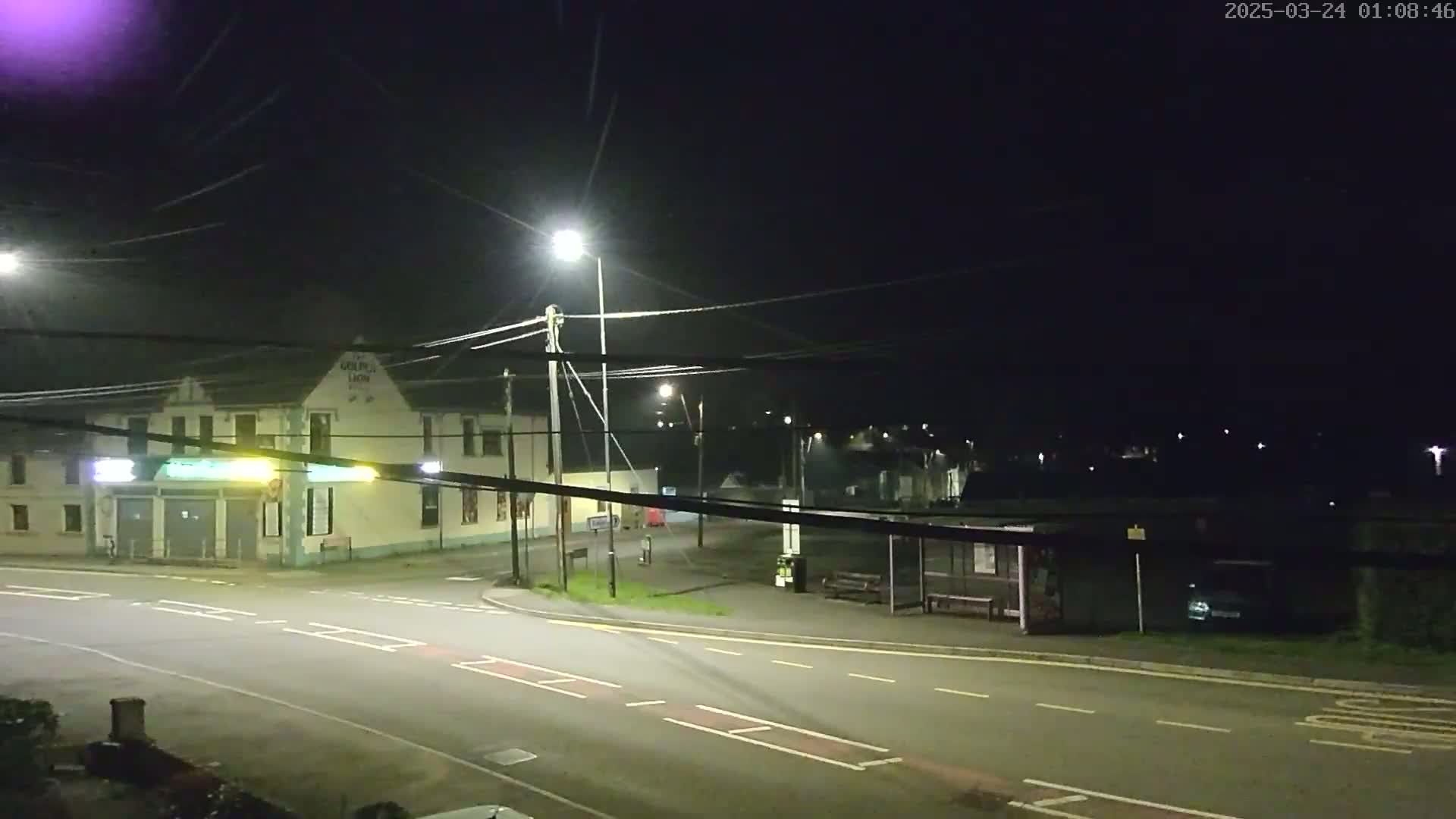 A nighttime street scene shows a building with illuminated signage, a bus stop, and streetlights illuminating a mostly empty intersection under a dark sky.