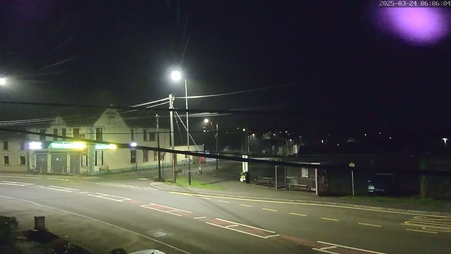 A nighttime street scene shows a crossroads with a building, a bus shelter, and streetlights illuminating the area under a clear, dark sky.