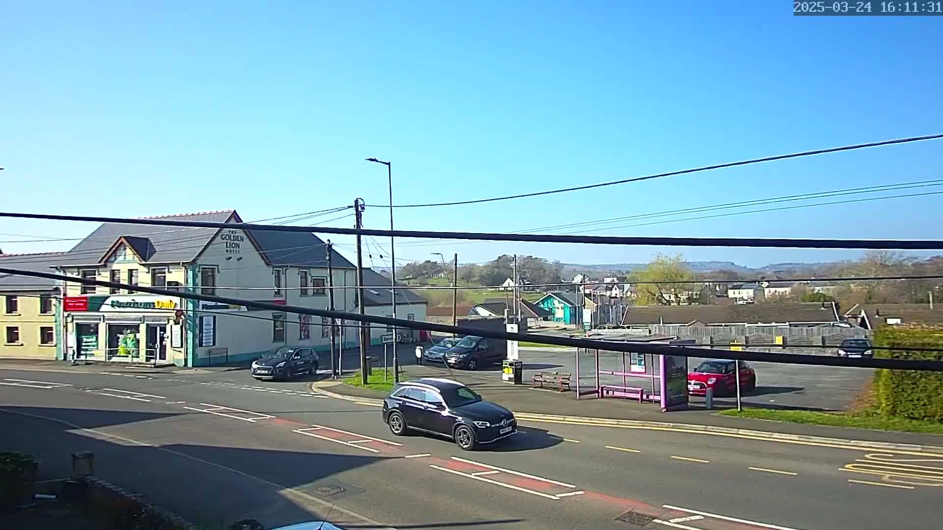 A black SUV drives past a corner intersection where a two-story building and a small convenience store are located, under a clear blue sky.