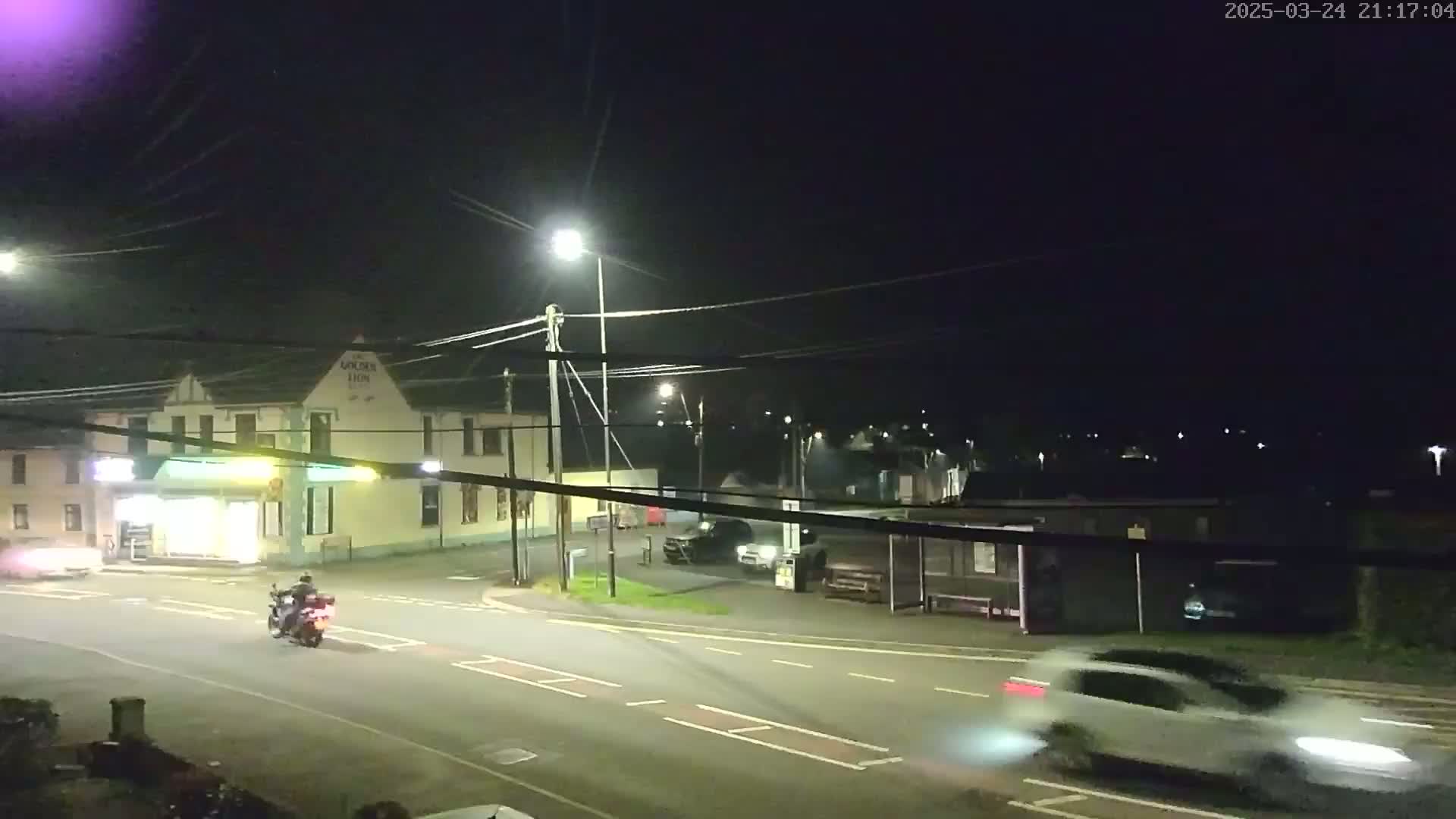 At night, a motorcycle and several cars are visible on a road near a building with streetlights illuminating the scene.