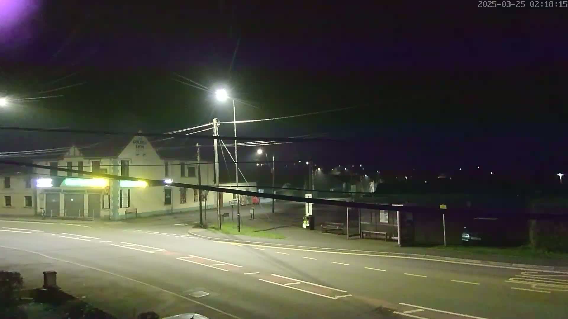 A nighttime view of a street intersection with a pub and bus stop, illuminated by streetlights under a dark sky.