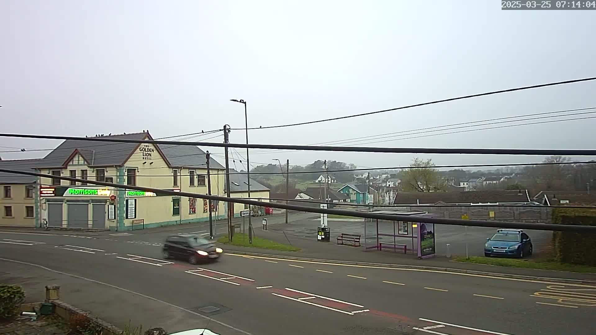 A grey overcast day shows a street scene with a pub, a bus stop, several cars, and houses on a hillside in the background.