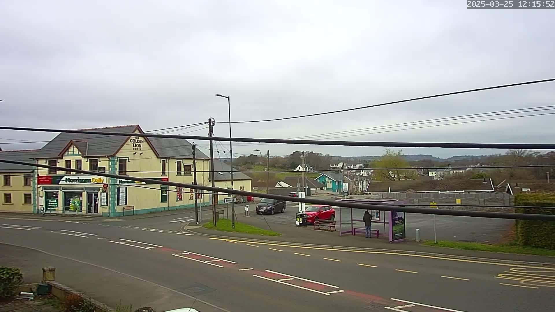 A view of a town intersection on an overcast day, showing a hotel, a convenience store, several cars, a bus stop with a person waiting, and a small residential area in the background.