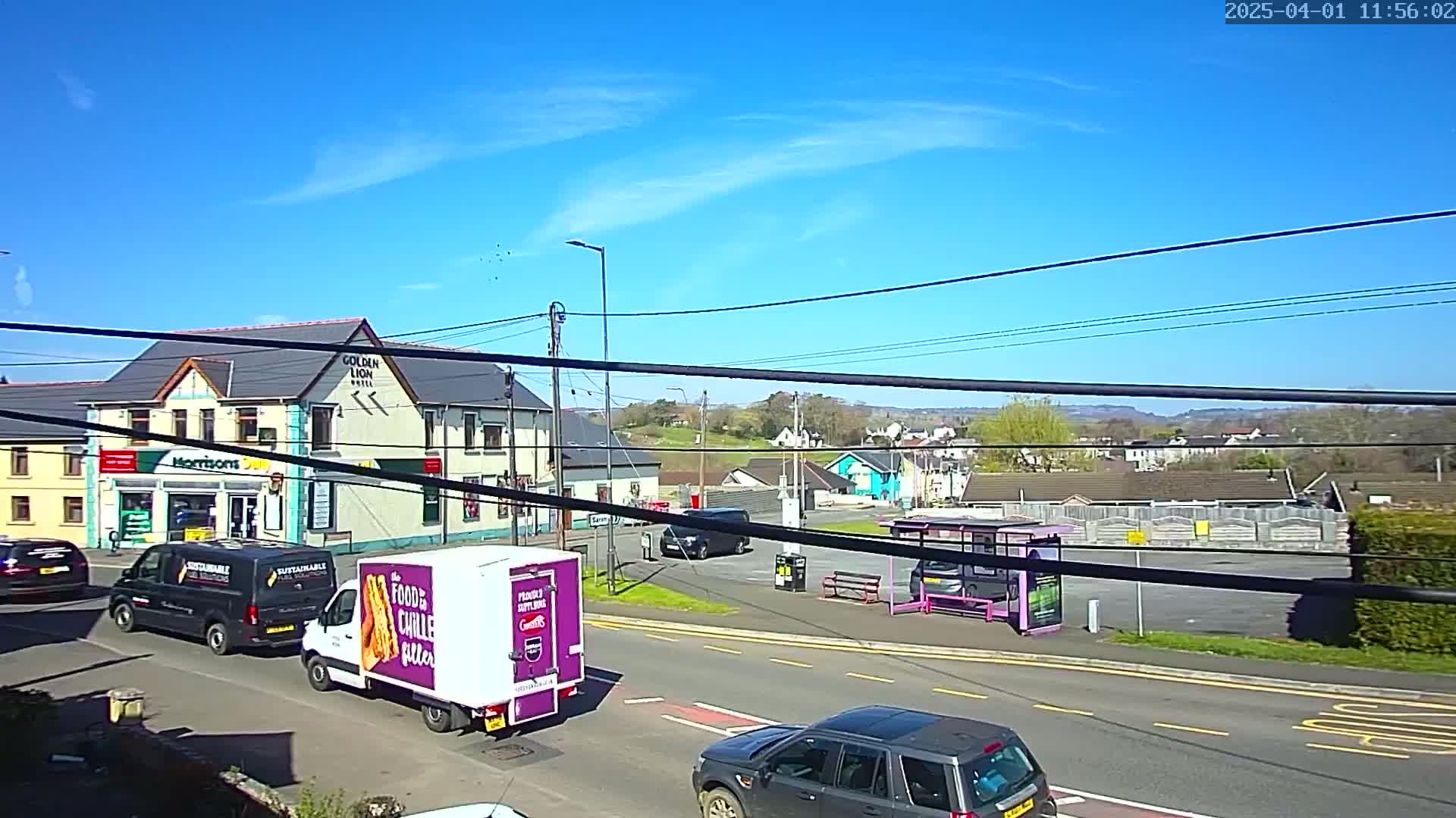 A sunny day reveals a street scene with several vehicles, including vans and a dark-colored SUV, near a building with a shop and a hotel, and a bus stop.
