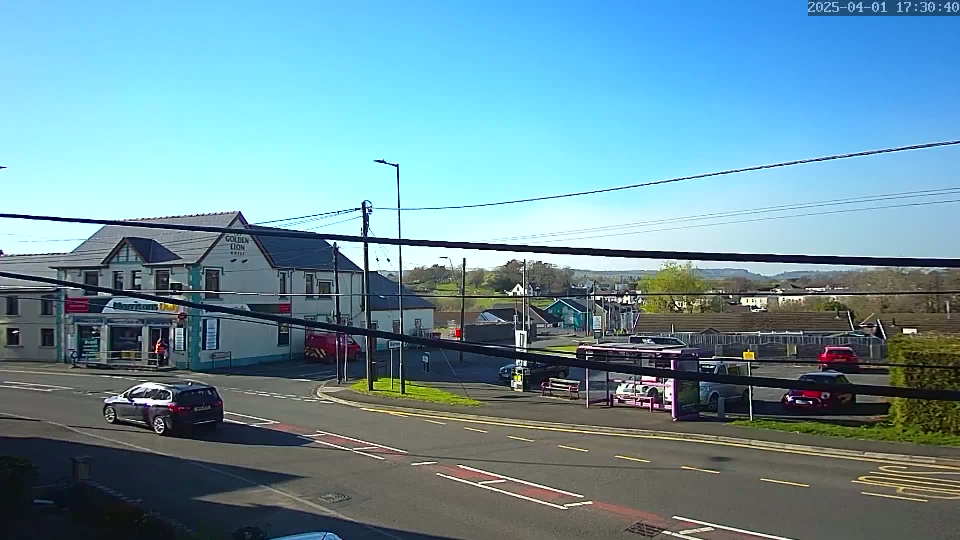 A sunny day reveals a street scene with a two-story building, several cars, a bus shelter, and a small parking lot; in the distance, hills and more buildings are visible under a clear blue sky.