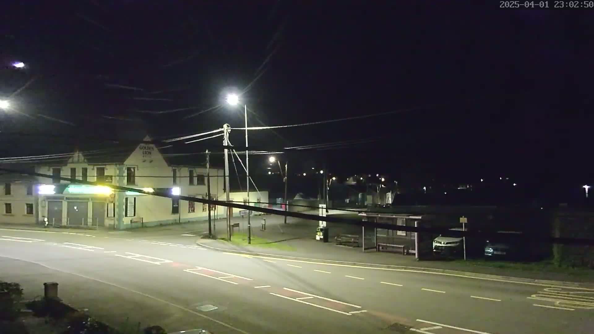 A nighttime street scene shows a building, streetlights, a bus stop with a couple of cars parked nearby, and an empty road under a clear, dark sky.