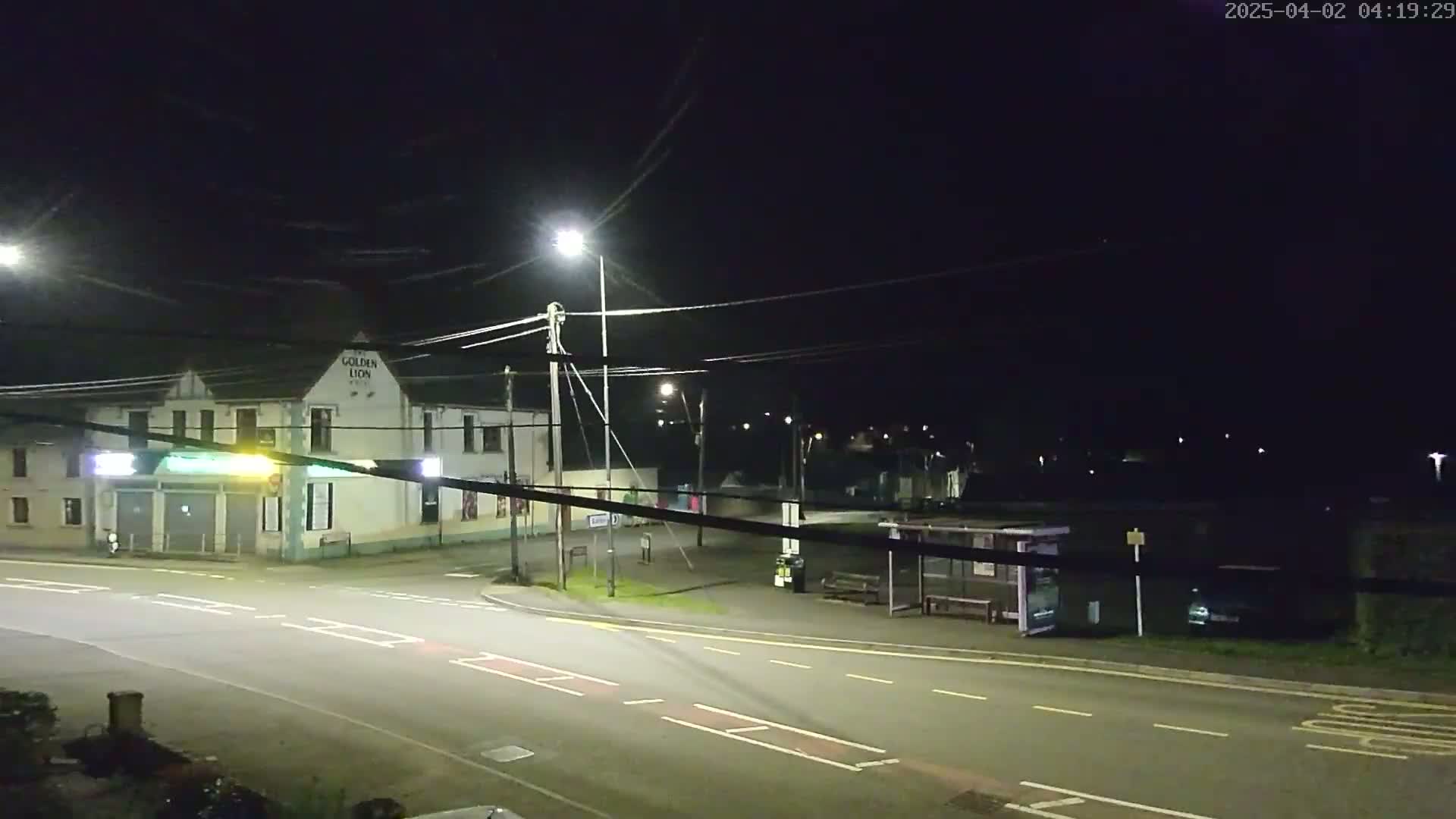 A nighttime street scene shows a corner intersection with a pub, a bus shelter, and several streetlights illuminating the road and surrounding area.