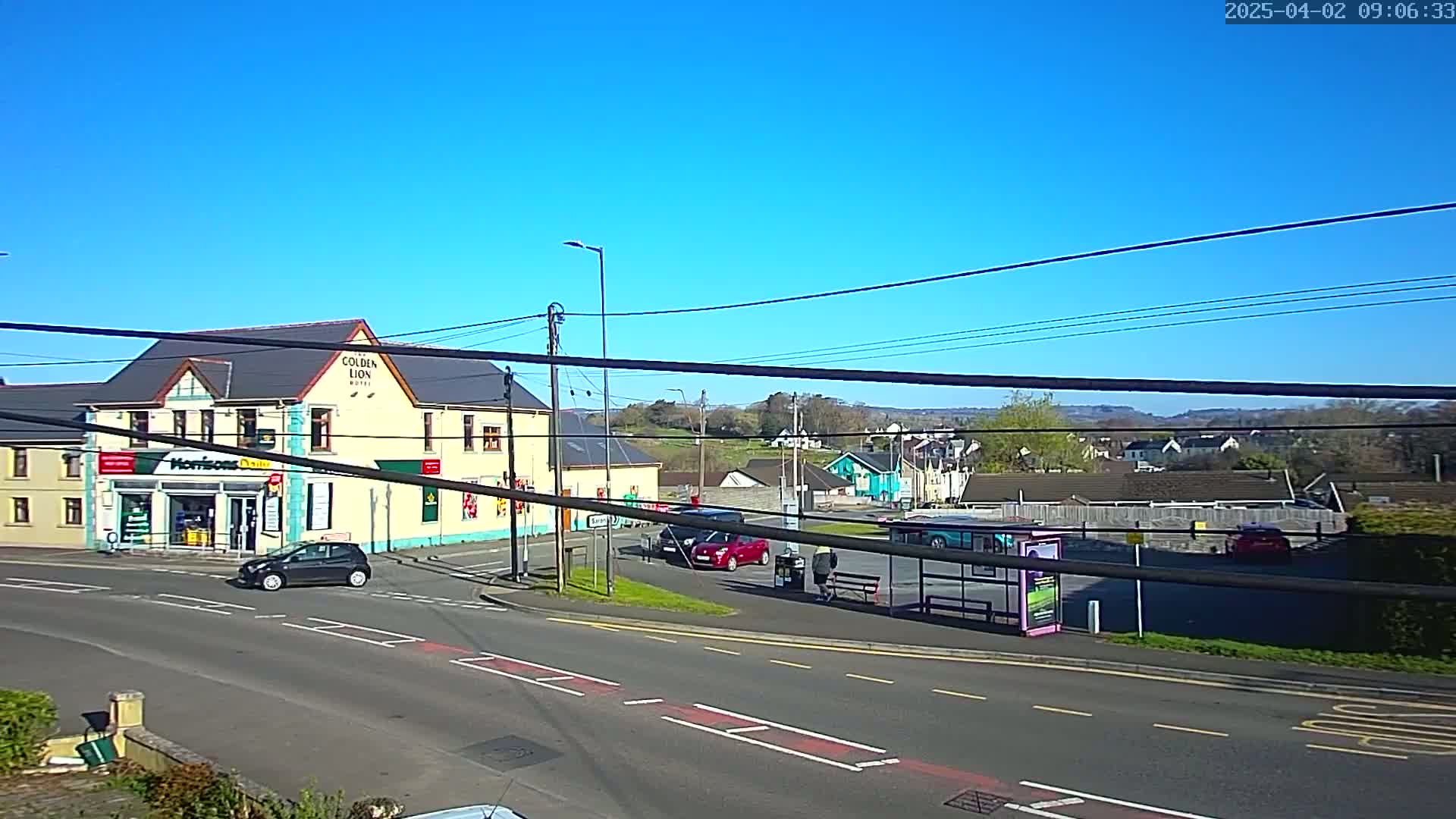 A sunny day reveals a town square with a hotel, a small shop, several cars, a bus stop, and a person standing near it, all under a clear blue sky.