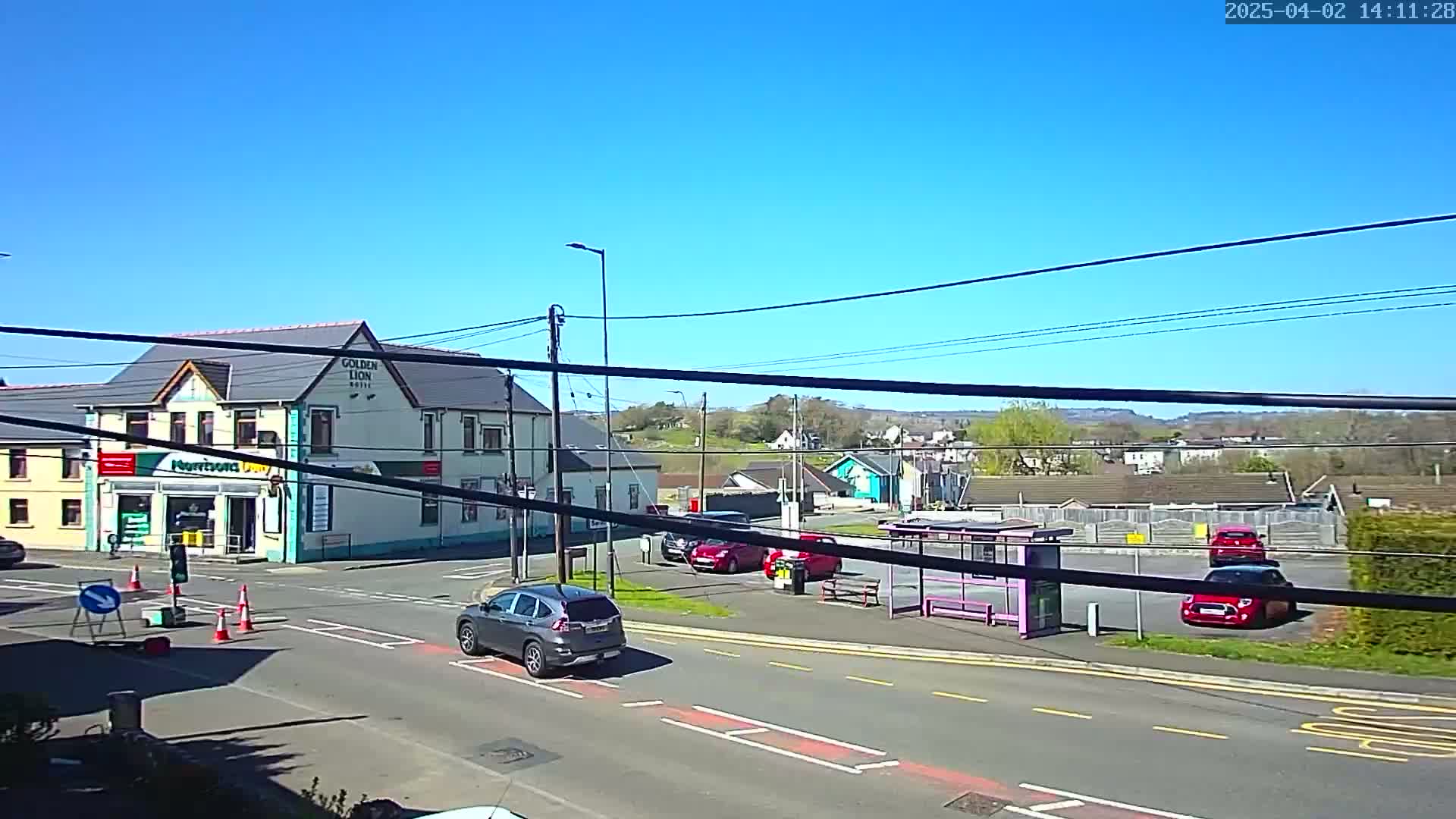 A sunny day shows a street scene with a two-story building, a bus stop, several cars, and a clear blue sky.