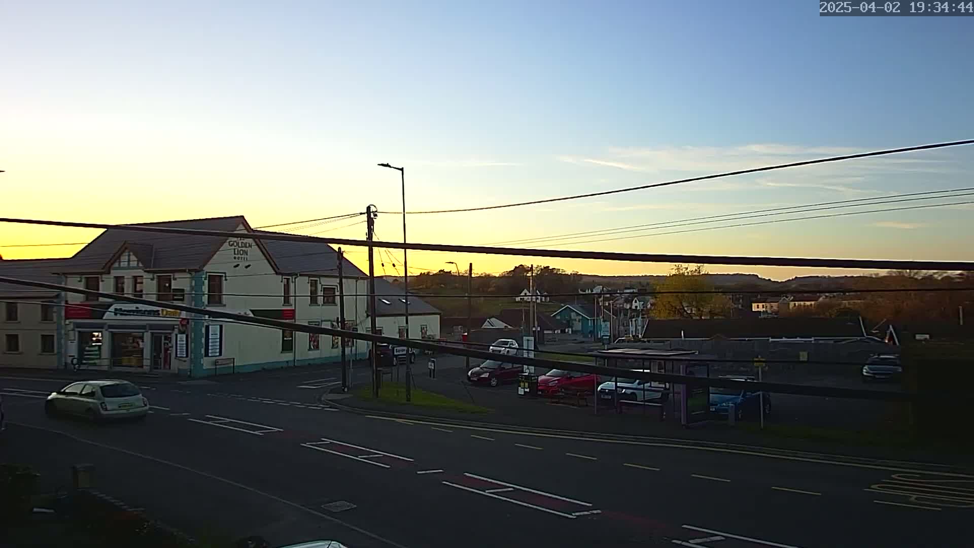 A street scene at sunset shows a two-story building, several cars parked in a lot, and a few cars driving on a road.