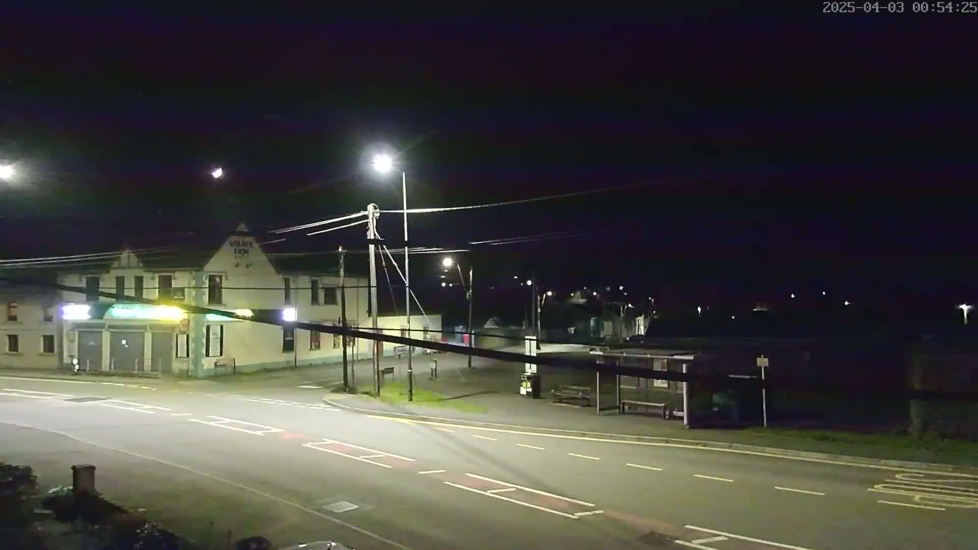 A nighttime street scene shows a pub, a bus shelter, and a road intersection illuminated by streetlights under a clear, dark sky.