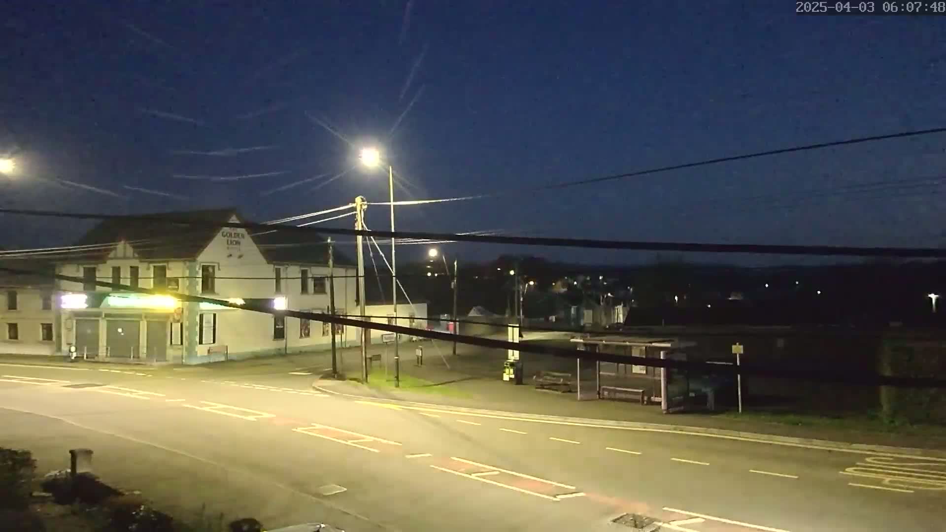 A nighttime, dimly lit street scene shows a corner building with a bus stop across the street, under a dark blue sky.