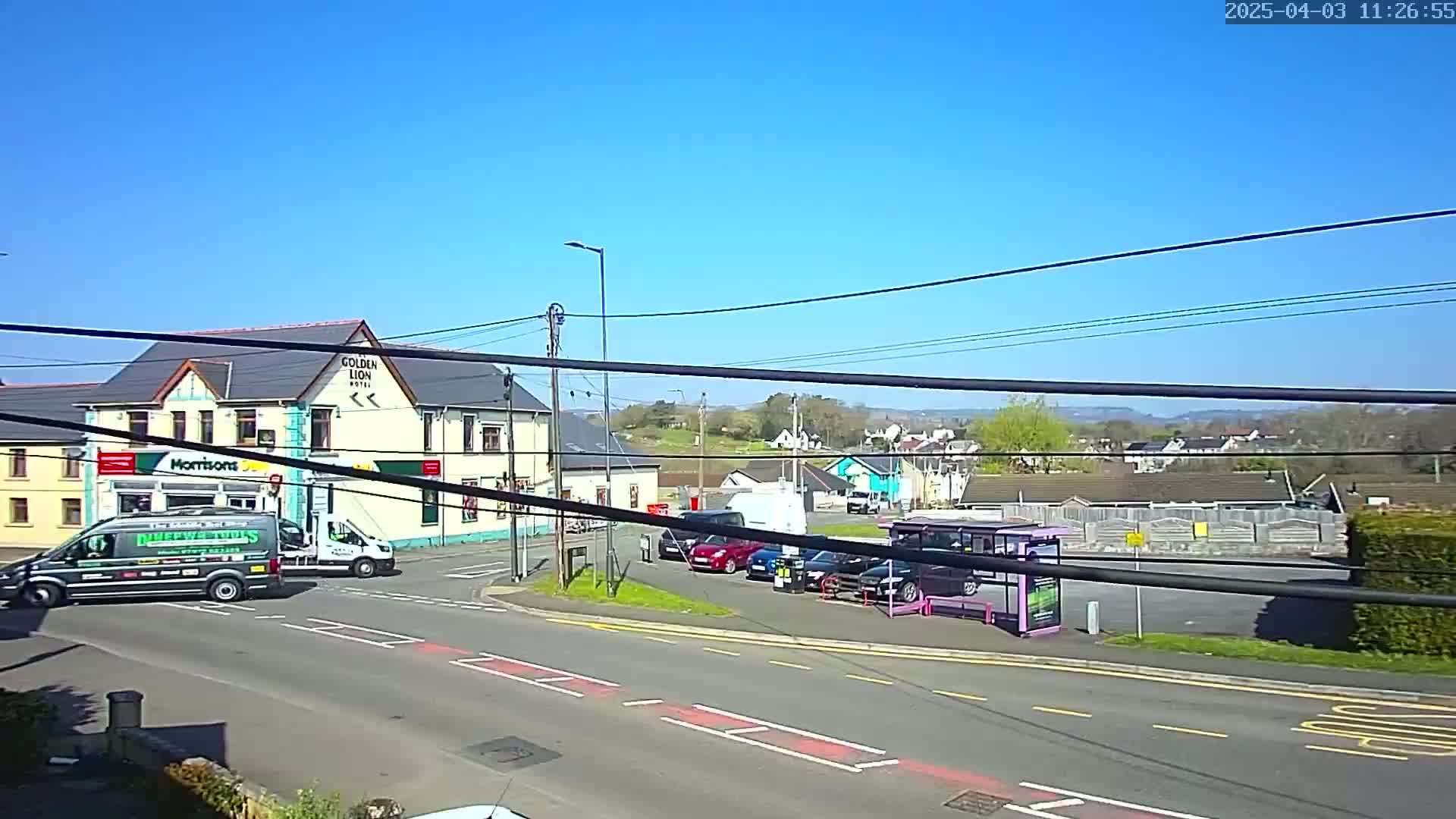 A sunny day shows a street scene with several vehicles, a hotel, and a bus stop, all situated near a small town with rolling hills in the background.