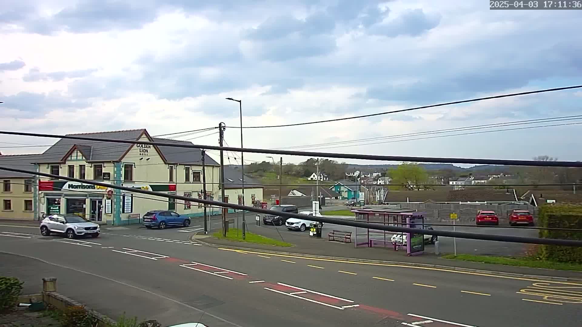 A partly cloudy day reveals a street scene with several cars parked near a bus stop and a pub, with additional buildings and houses visible in the background.