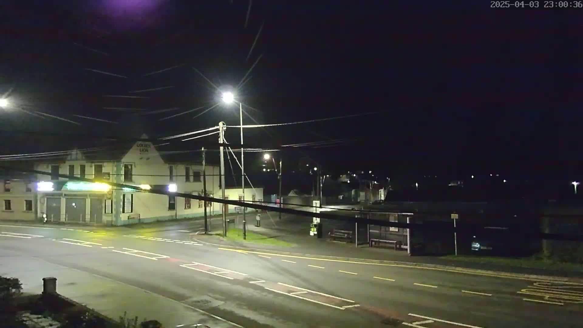 A nighttime street scene shows a building with lights, streetlights, a bus shelter, and a sparsely trafficked road under a dark, clear sky.