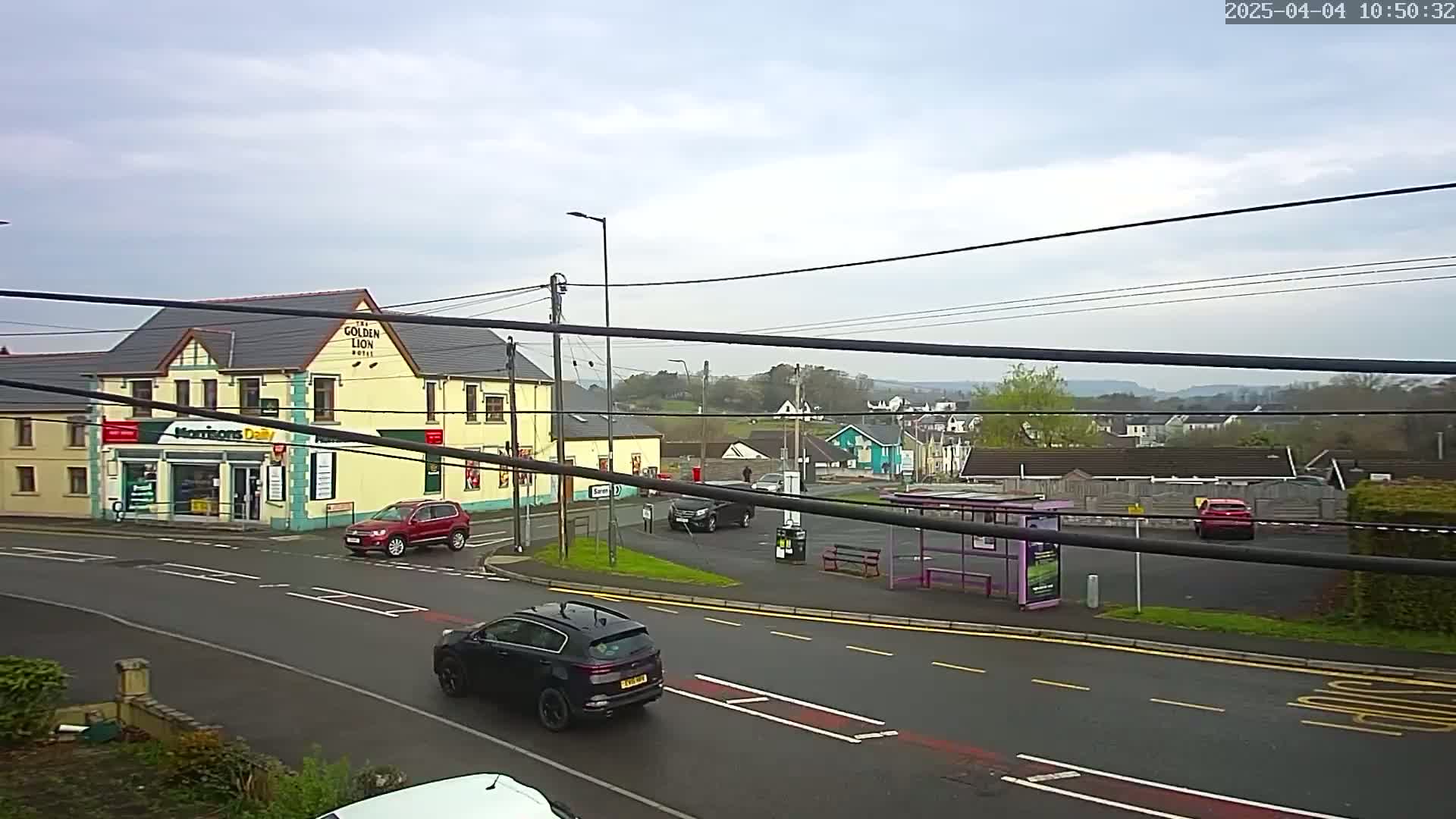 A street scene on an overcast day shows several cars parked and driving around a small town square with a pub, a shop, and a bus stop.