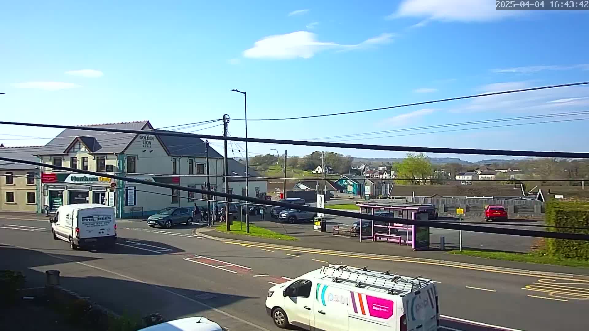 A sunny day reveals a street scene with several vehicles, including vans and cars, near a two-story building and a bus stop, with a background of trees and distant hills.