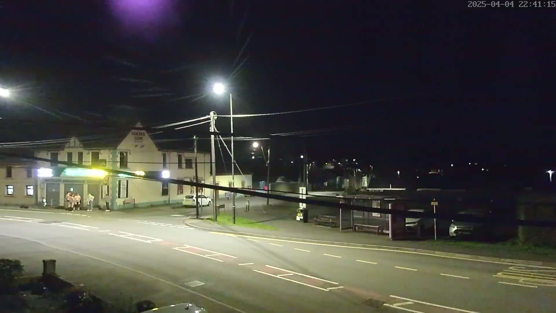 A nighttime street scene shows a pub, several parked cars, a bus shelter, and a few pedestrians standing near the pub under bright streetlights.