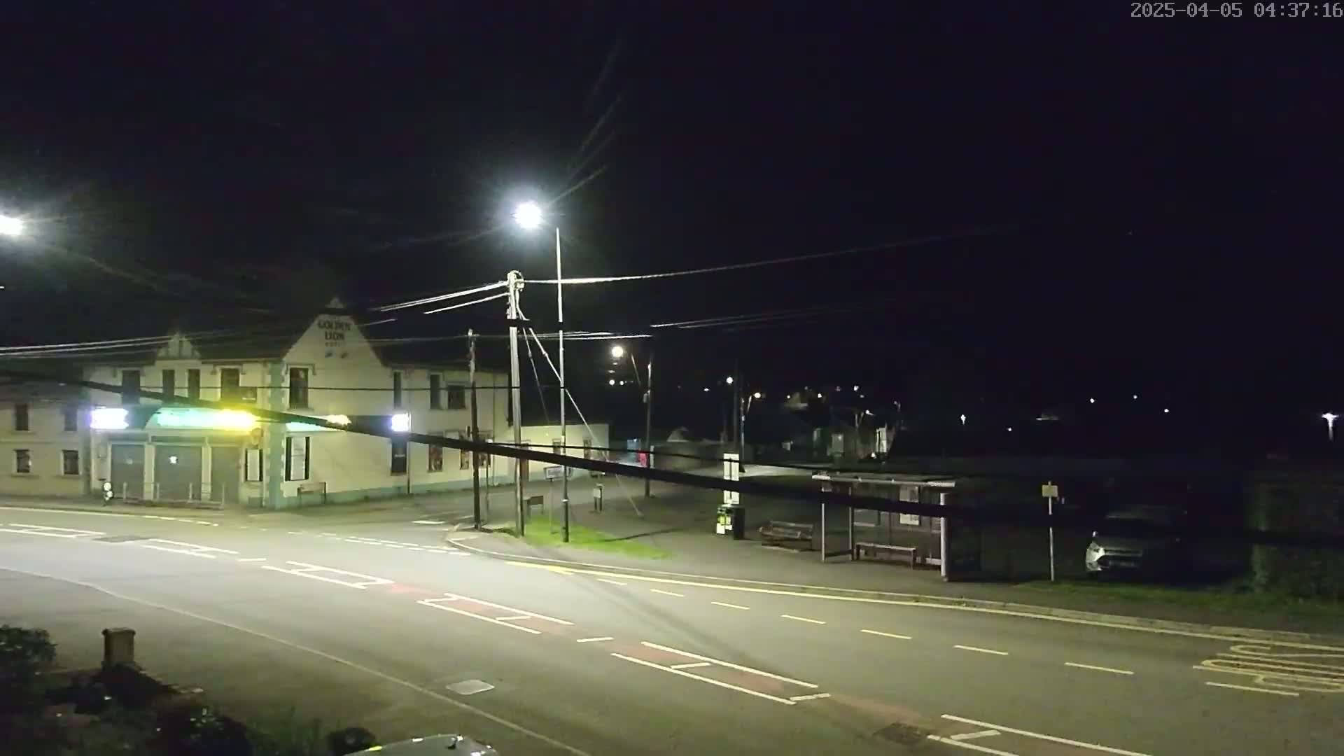 A nighttime street scene shows a mostly empty road intersecting in front of a building with illuminated signage, a bus stop, and a few parked cars under artificial lighting.
